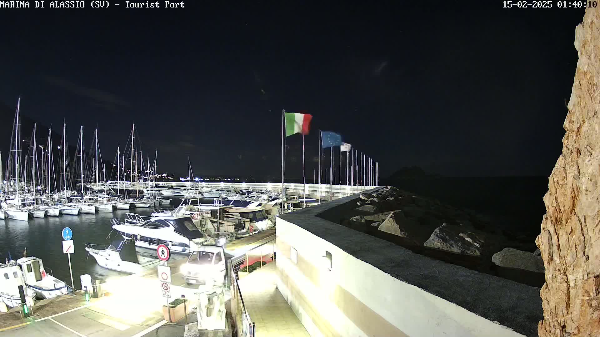 A nighttime view of a marina filled with sailboats and motorboats, under a clear, dark sky, with several flags waving near a rocky shoreline.