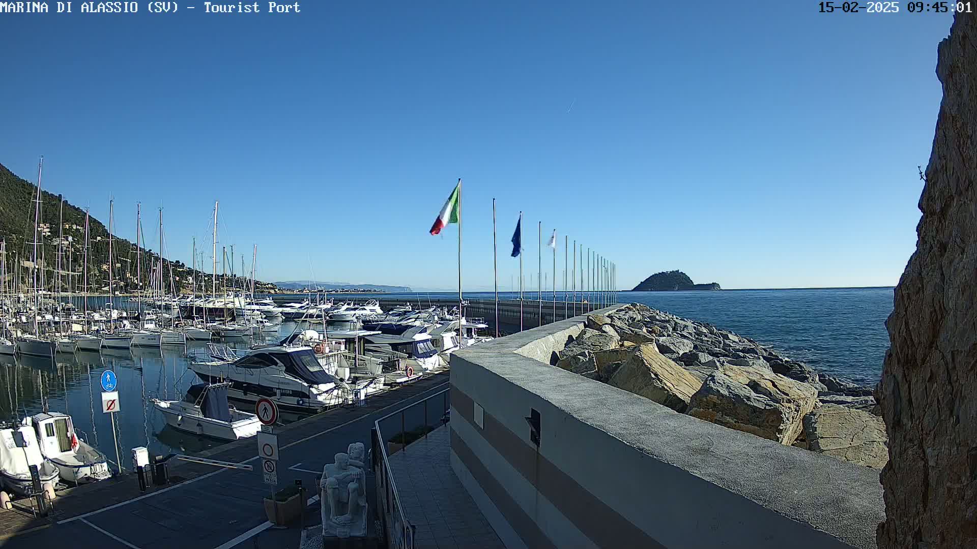 A marina filled with boats is seen on a sunny day, with a rocky seawall separating it from the calm blue sea and a small island visible in the distance.