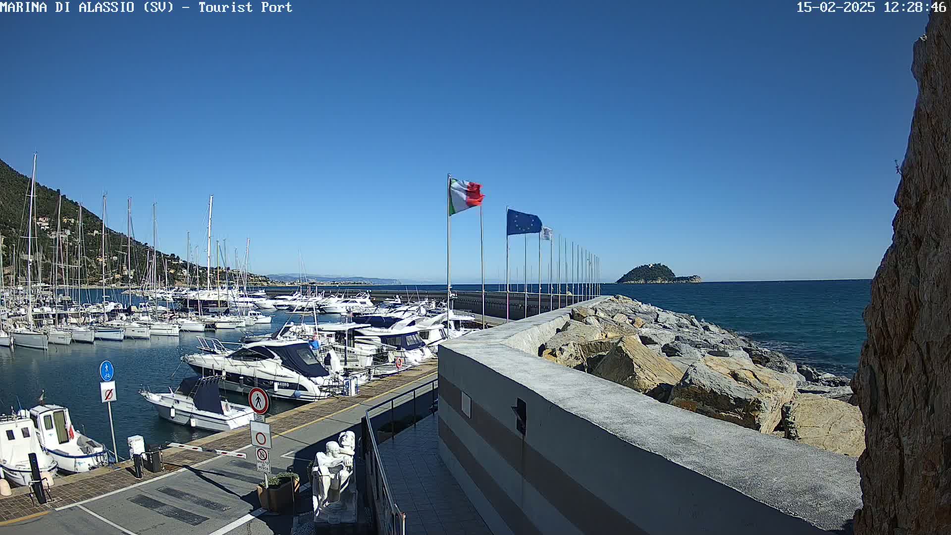 A marina filled with sailboats and motorboats is situated next to a rocky breakwater on a clear, sunny day.