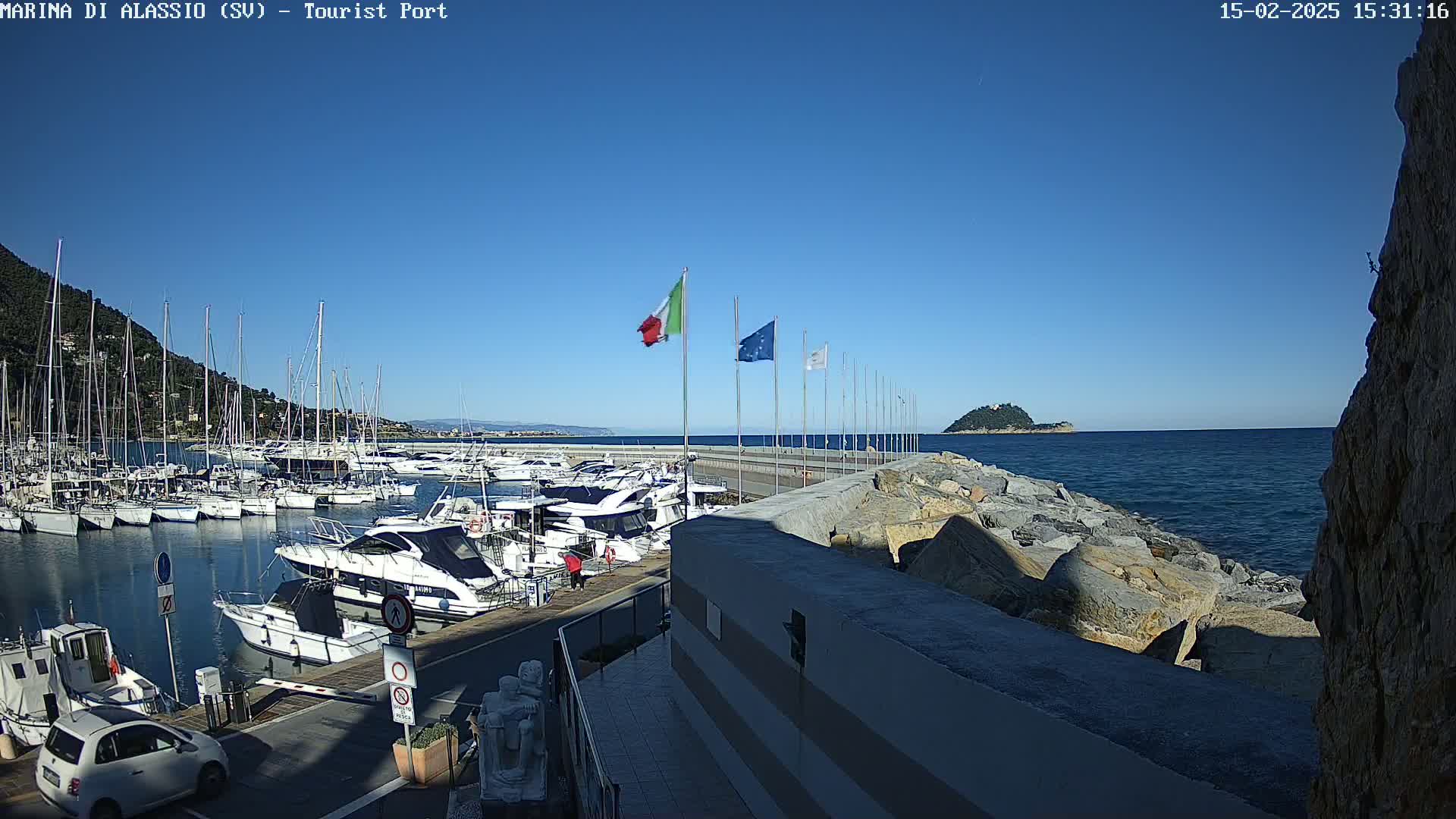 A marina filled with boats is situated next to a rocky shoreline on a clear, sunny day.