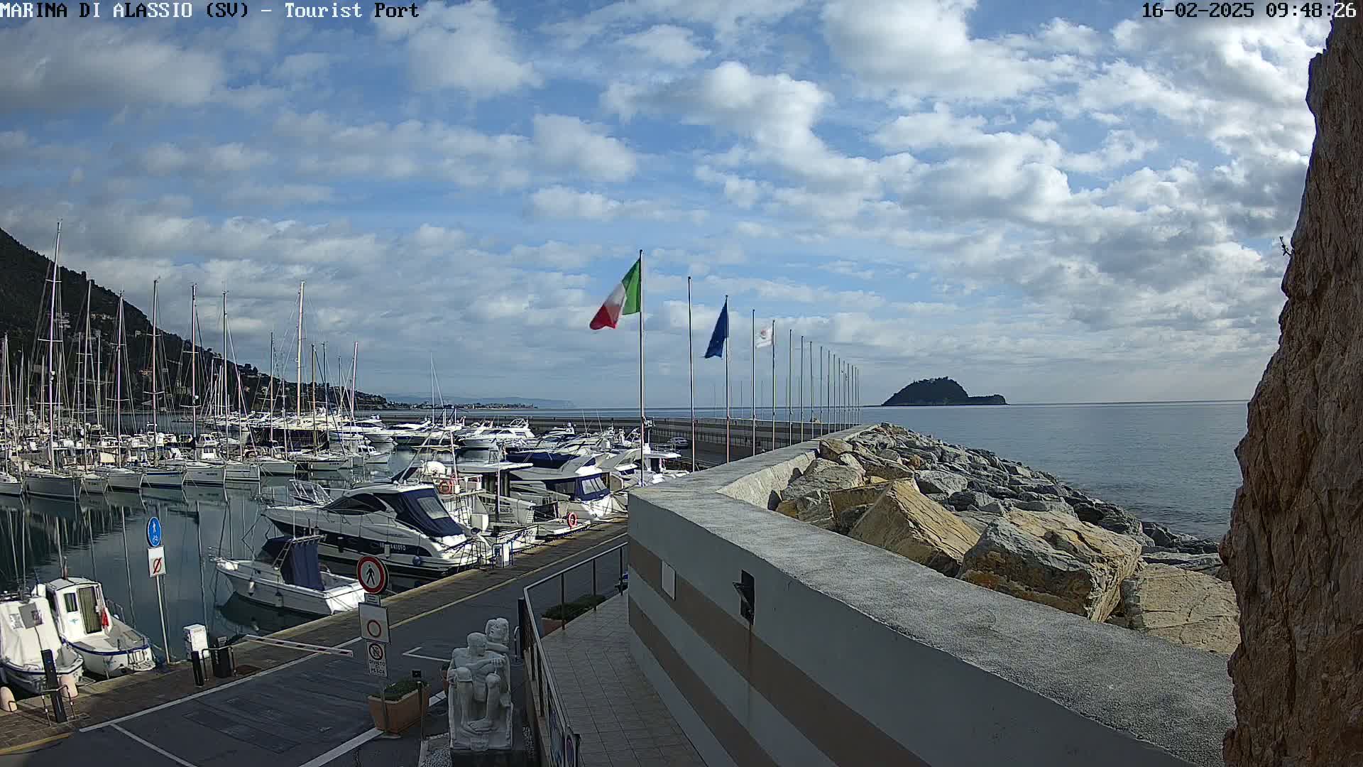 A marina filled with boats is situated next to a rocky seawall on a partly cloudy day.
