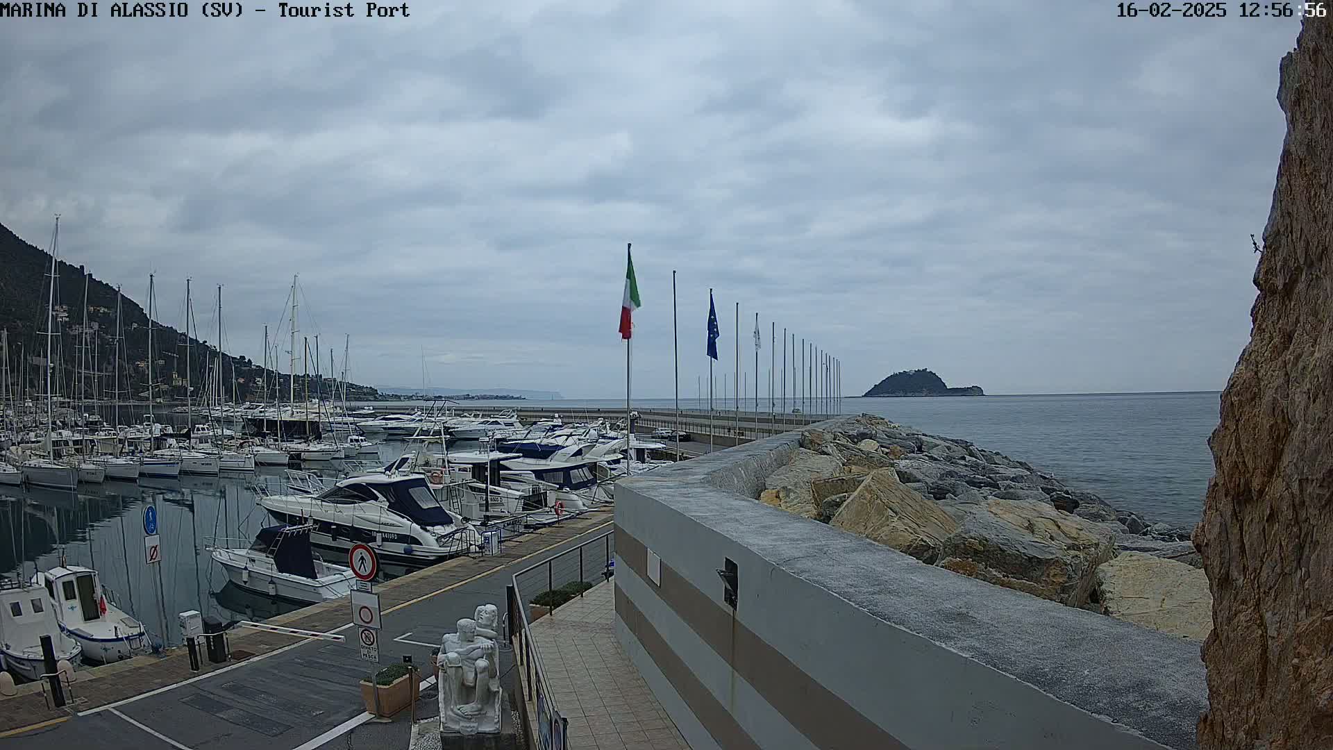 A marina filled with sailboats and motorboats is situated on a calm sea under an overcast sky, with a rocky embankment and distant hills visible.