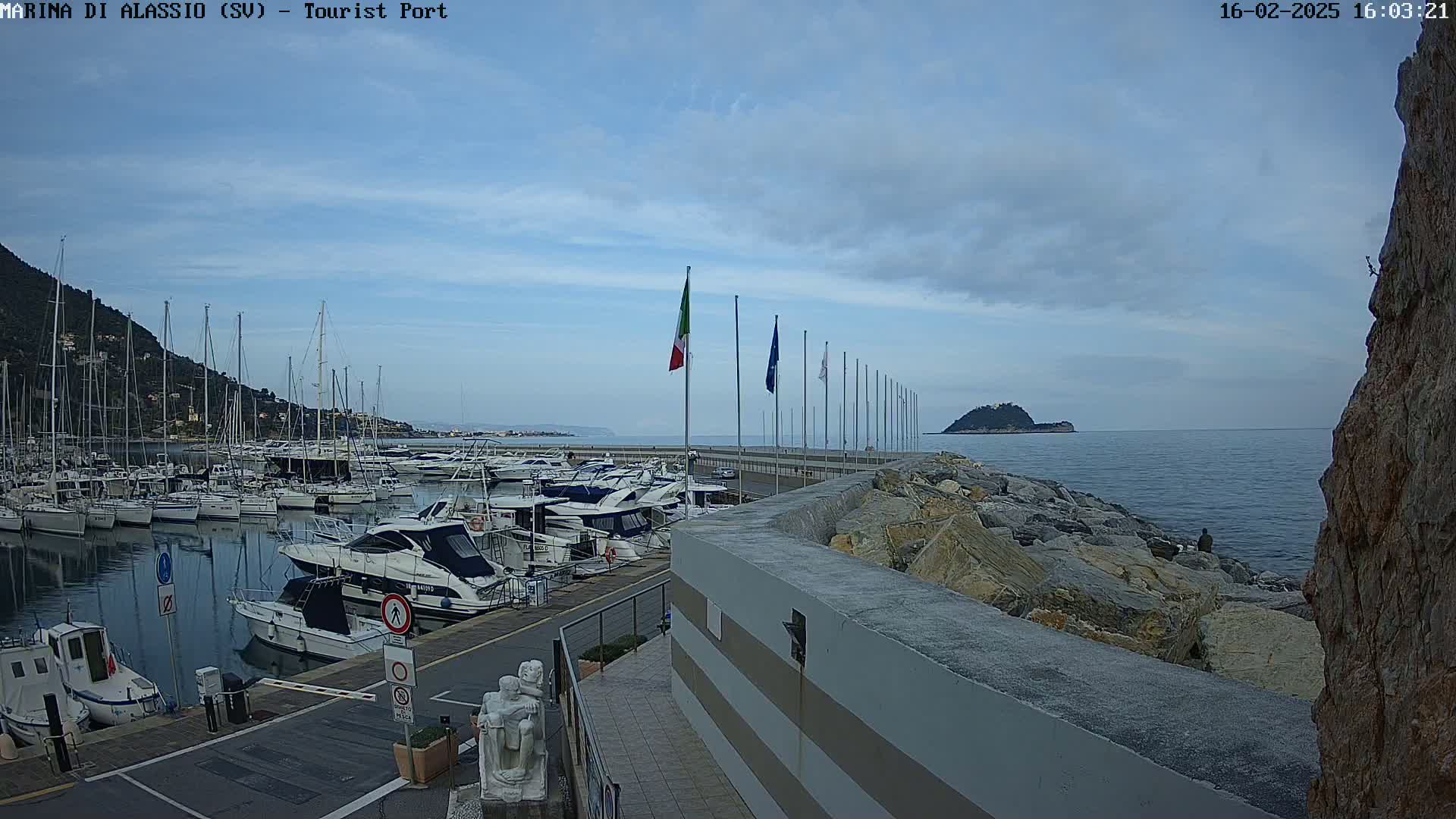 A marina filled with sailboats and motorboats is situated next to a rocky seawall under a partly cloudy sky.