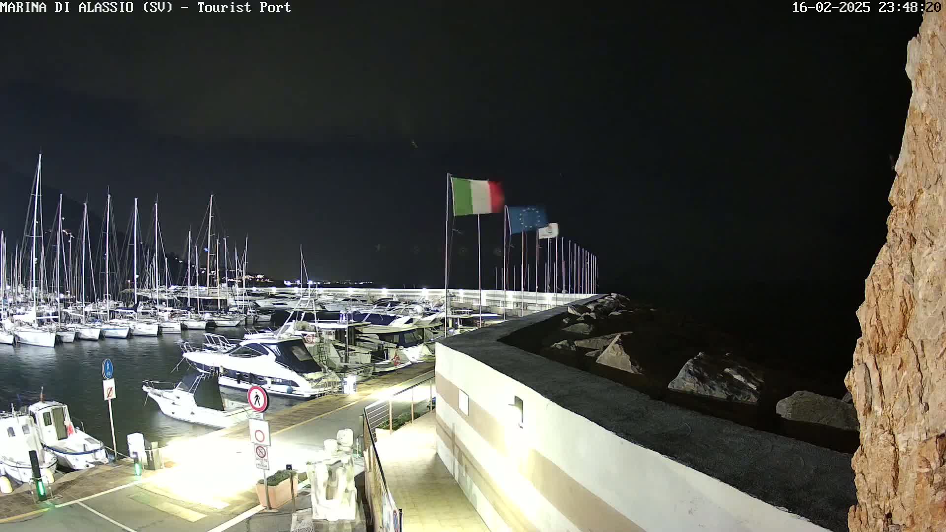 A nighttime view of a marina filled with sailboats and motorboats, under a clear, dark sky, with Italian and European Union flags waving on flagpoles.