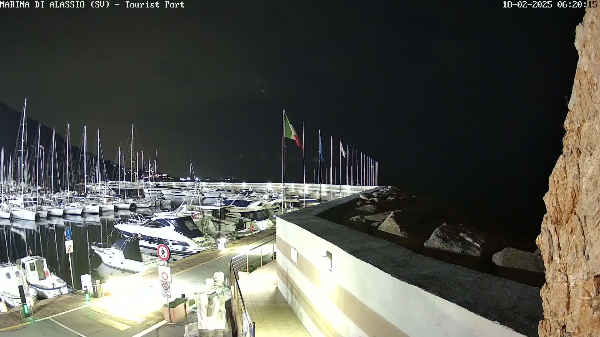 A nighttime view of a marina filled with boats, with an Italian flag flying and a rocky outcrop to the right under a clear, dark sky.