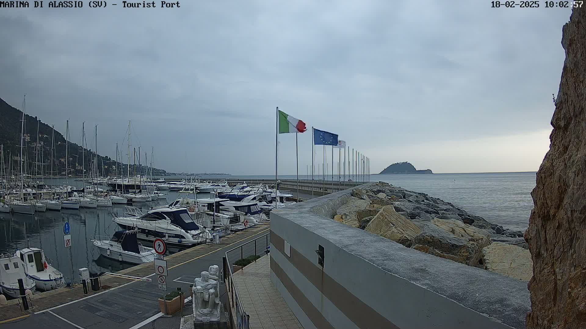 A harbor filled with numerous boats is situated next to a rocky shoreline under an overcast sky.