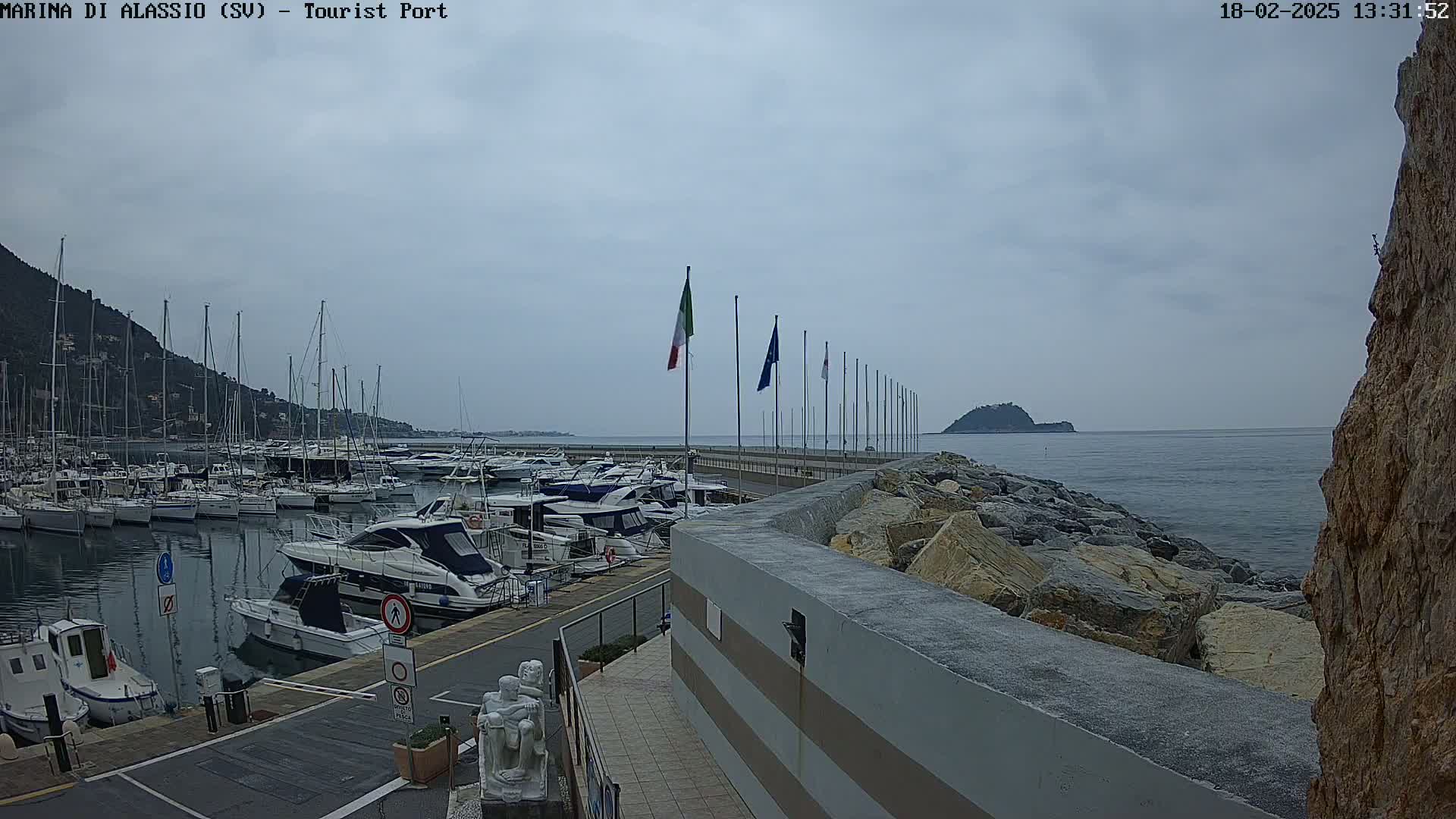 A marina filled with numerous boats is shown on an overcast day, with calm water extending to a small island in the distance.