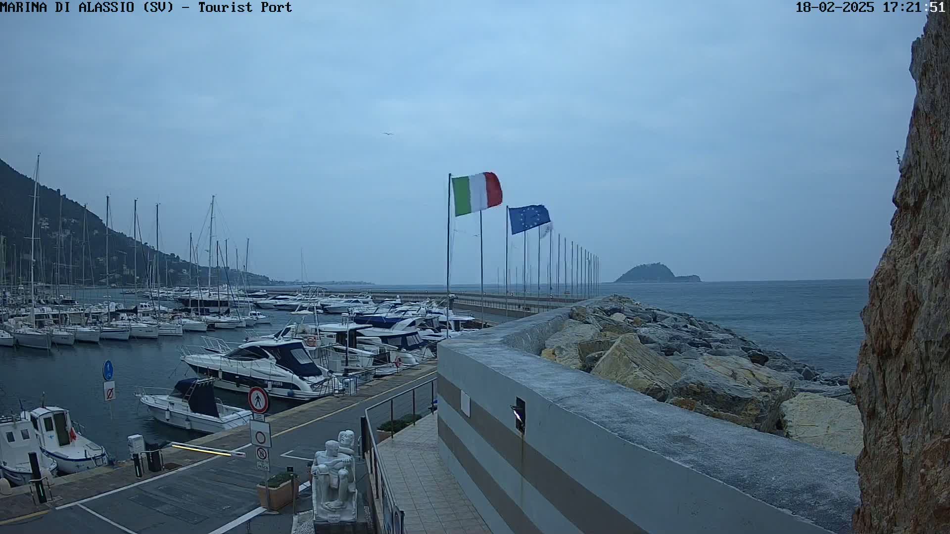 A harbor filled with boats is shown under an overcast sky, with Italian and European Union flags flying near a rocky breakwater.