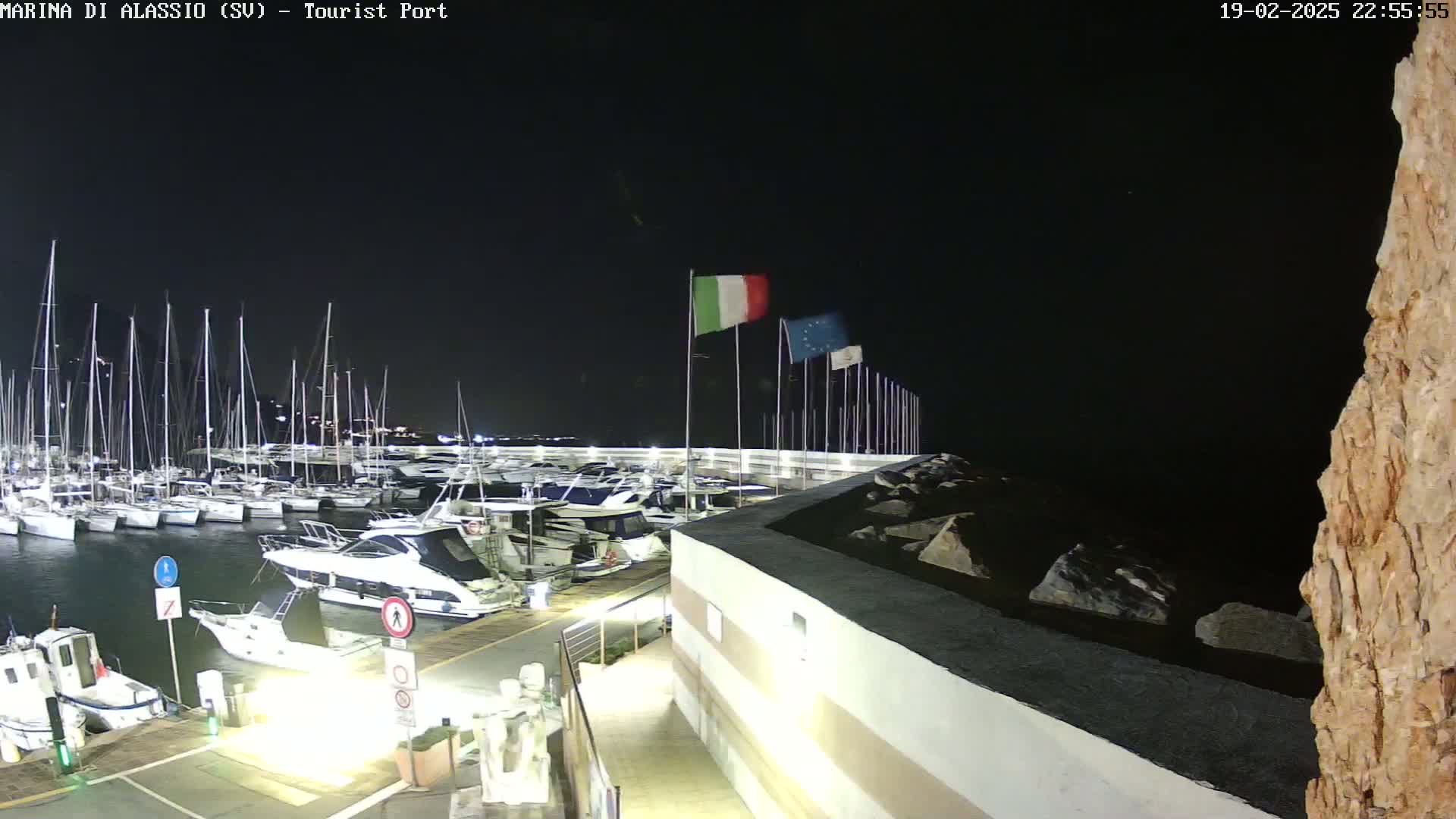 A nighttime view of a marina filled with sailboats and motorboats, with Italian and European Union flags waving in the dark sky.