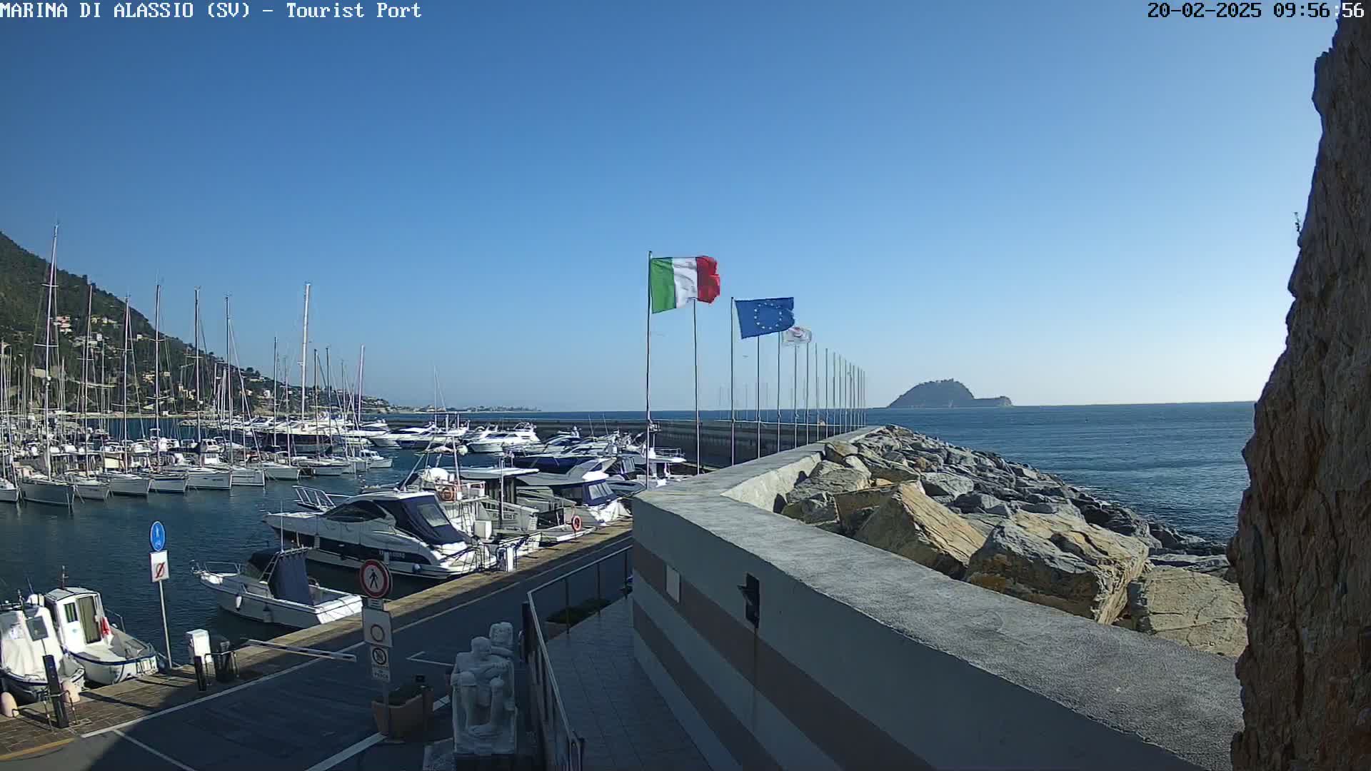 A marina filled with sailboats and motorboats is situated beside a rocky wall overlooking a calm sea under a clear blue sky.