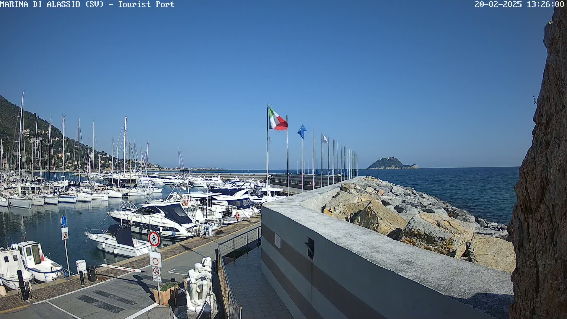 A marina filled with boats is situated on a calm, sunny day next to a rocky shore and a small island in the distance.