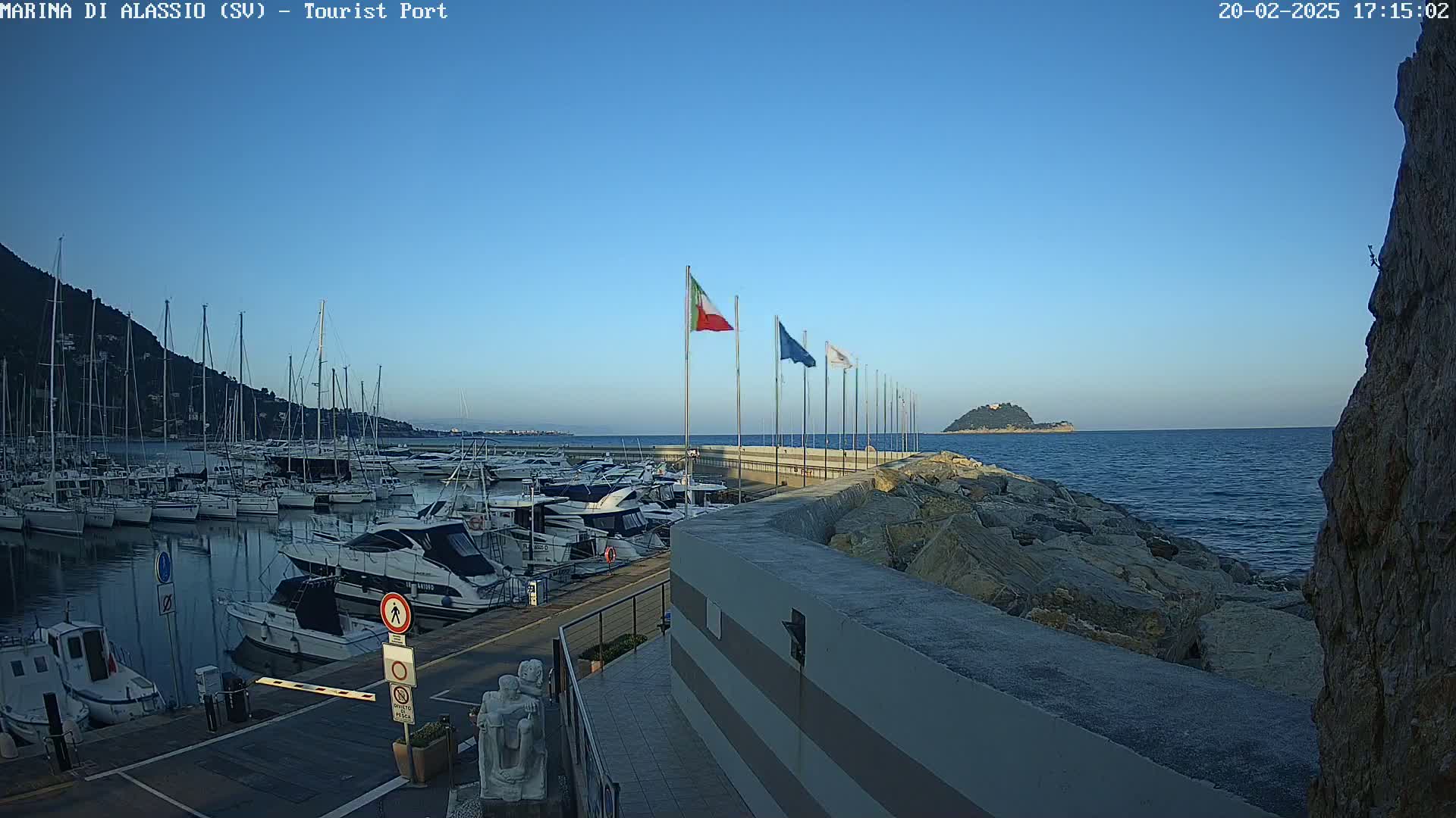 A marina filled with boats is situated next to a rocky shoreline, with a clear blue sky and calm sea in the background.