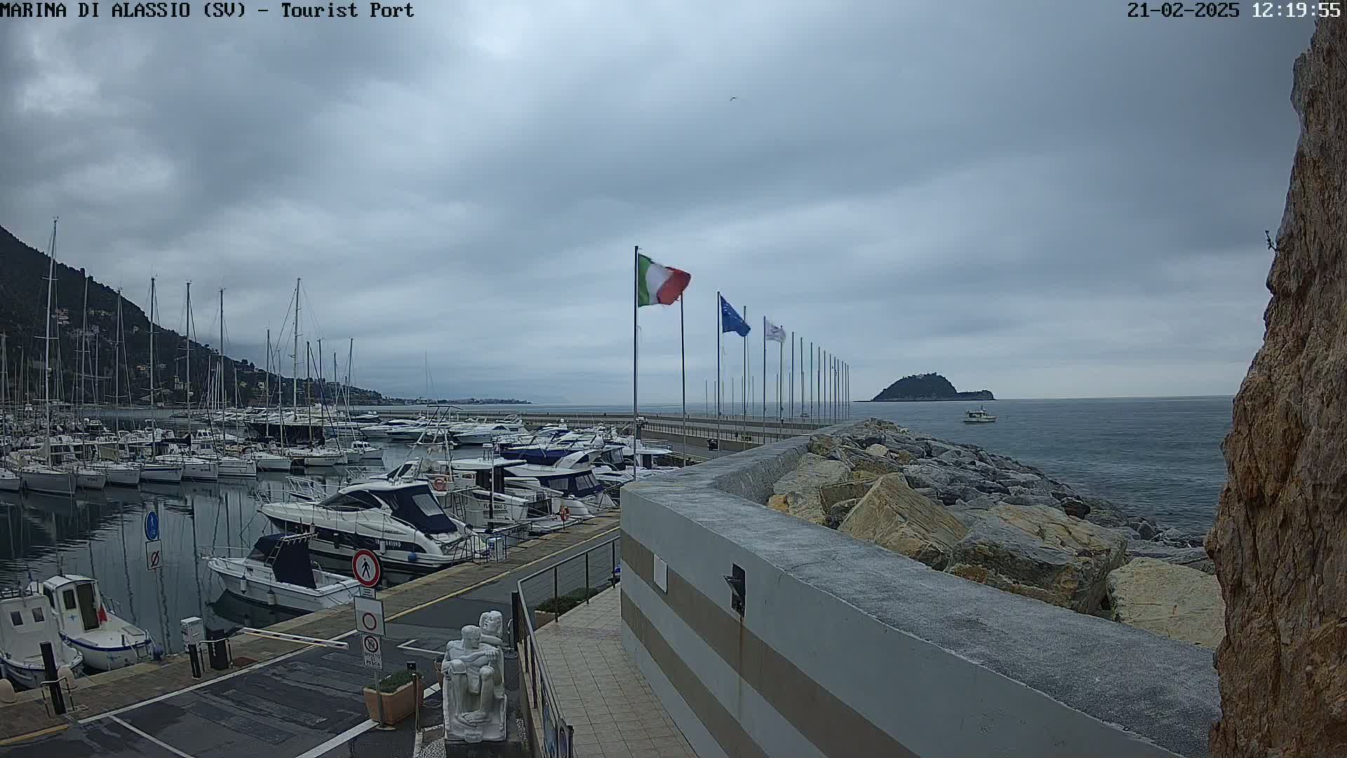 A harbor filled with sailboats and motorboats under a cloudy sky, with a rocky breakwater and a small island in the distance.