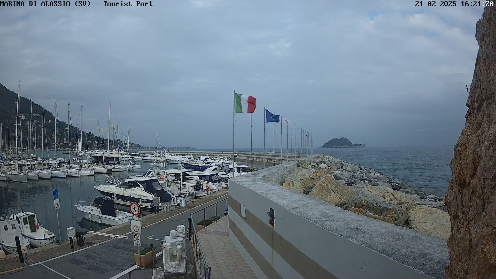 A marina filled with boats is situated next to a rocky shoreline under a cloudy sky, with several flags including the Italian flag flying above.