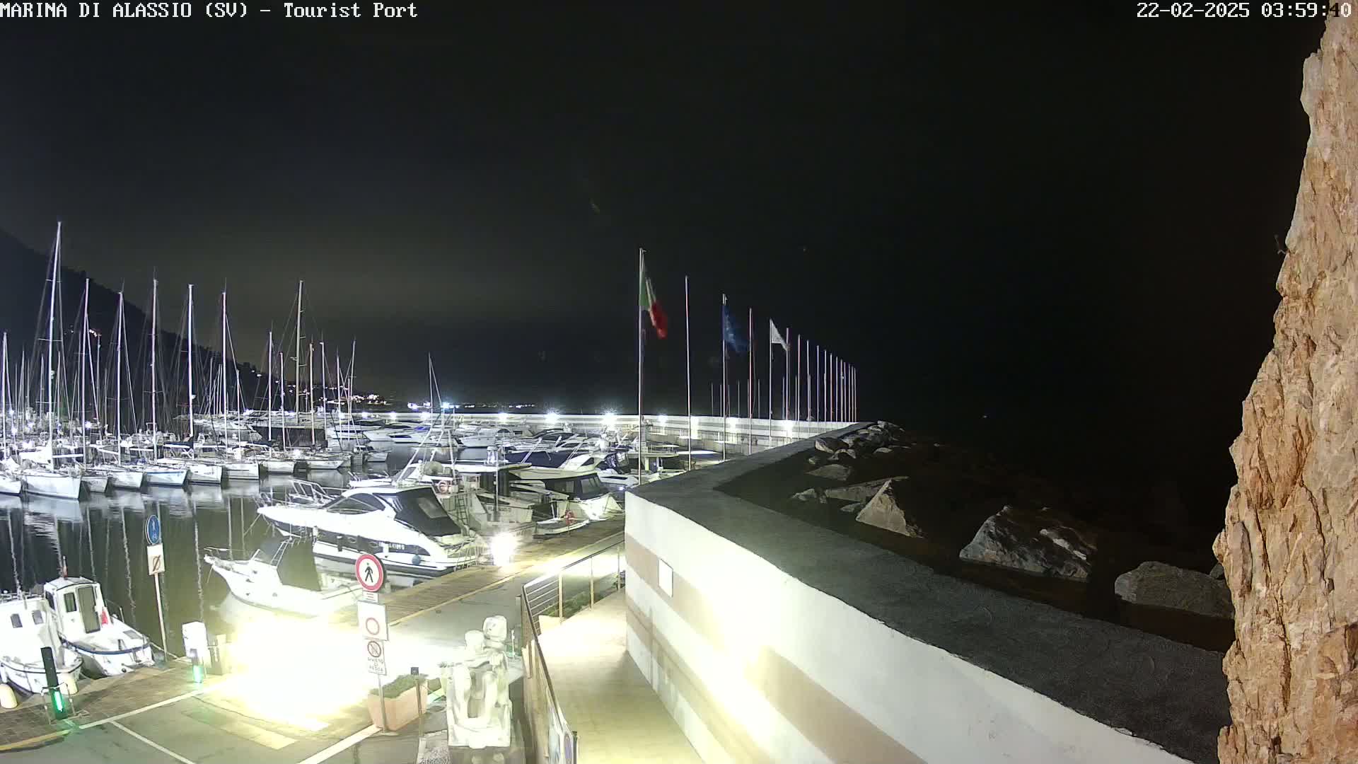 A nighttime view of a marina filled with boats, docked alongside a lit walkway and a rocky shoreline under a dark sky.