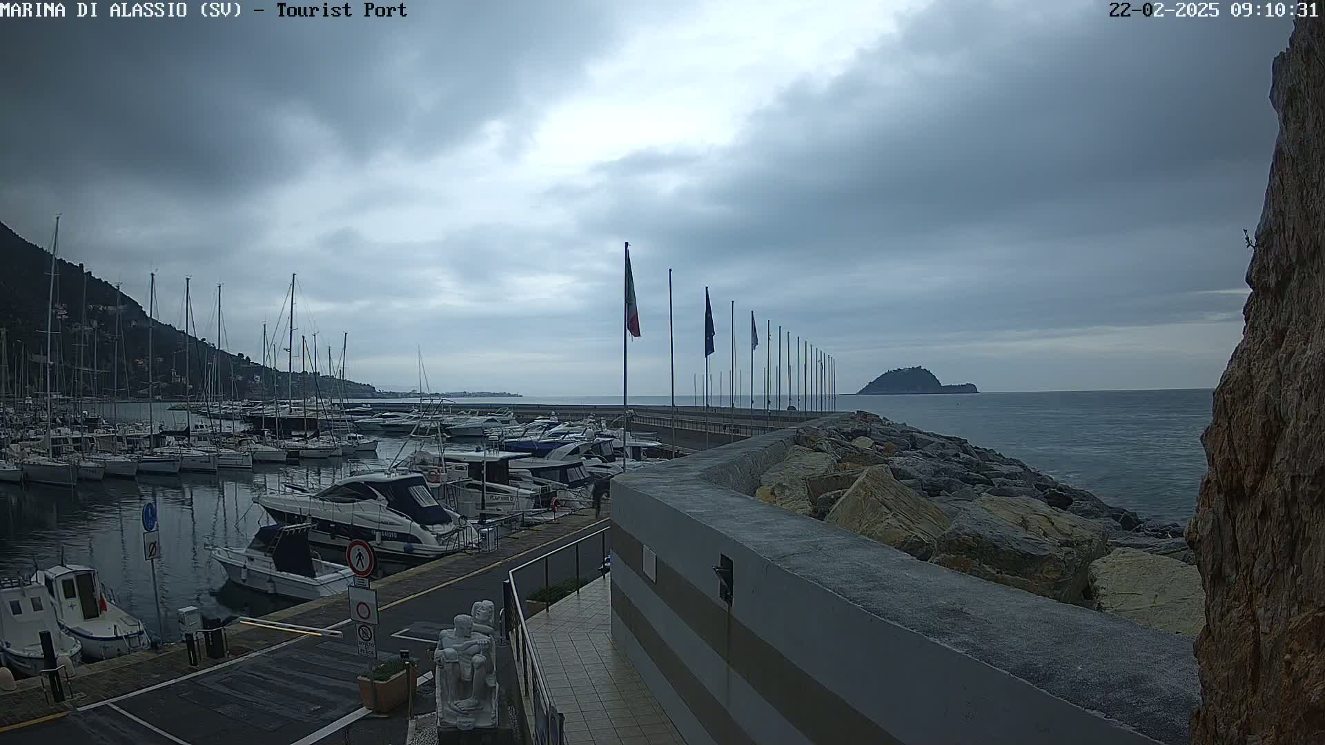 A harbor filled with boats is seen under an overcast sky, with a rocky breakwater separating it from the open sea and a small island in the distance.