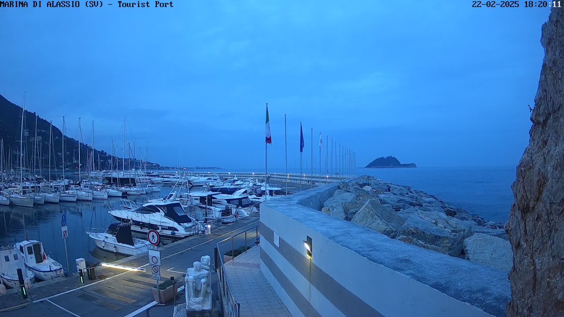 Under a twilight blue sky, a marina filled with boats is situated next to a rocky shoreline with a small island visible in the distance.