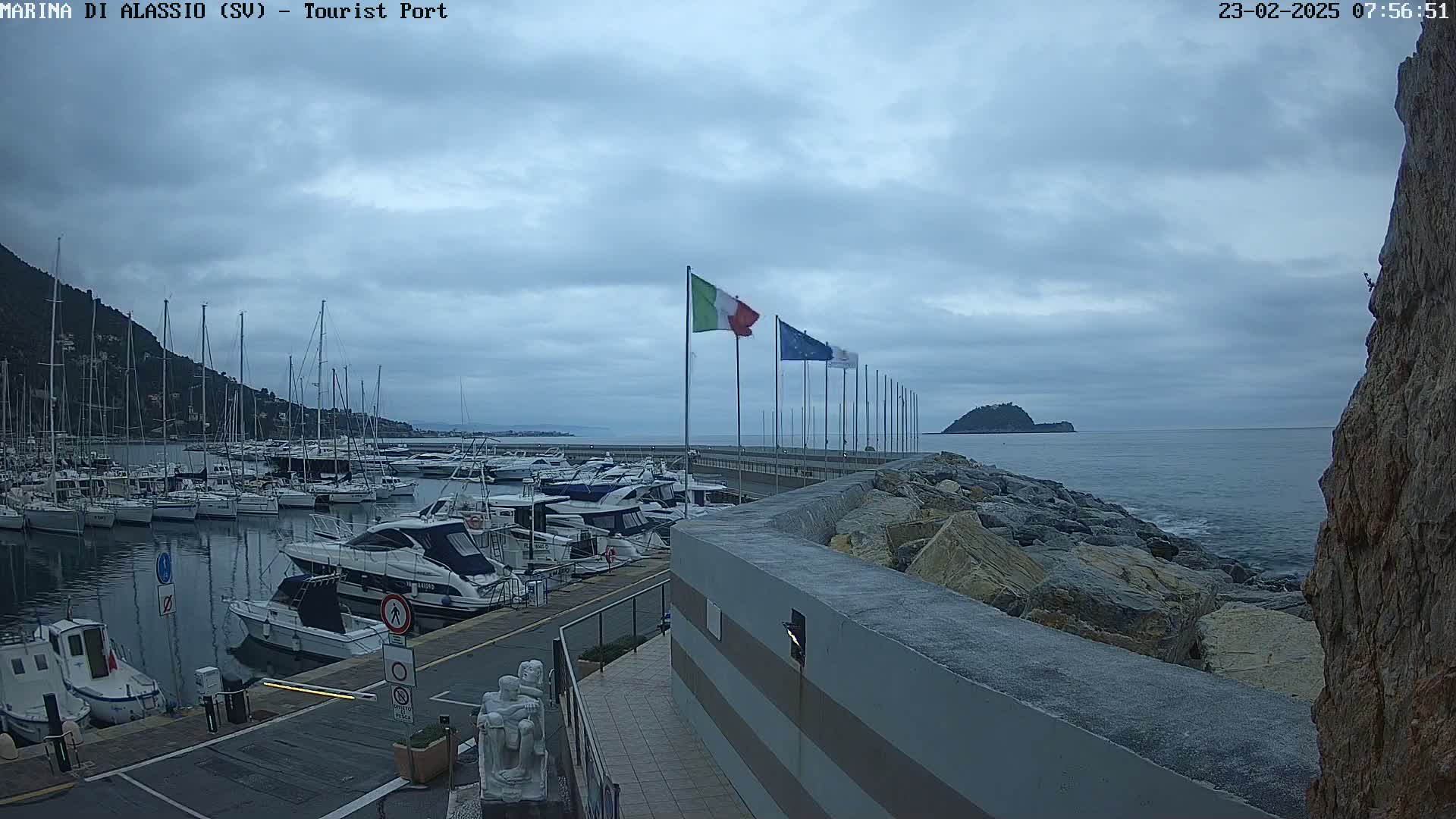 A marina filled with sailboats and motorboats is situated next to a rocky shoreline under a cloudy sky.