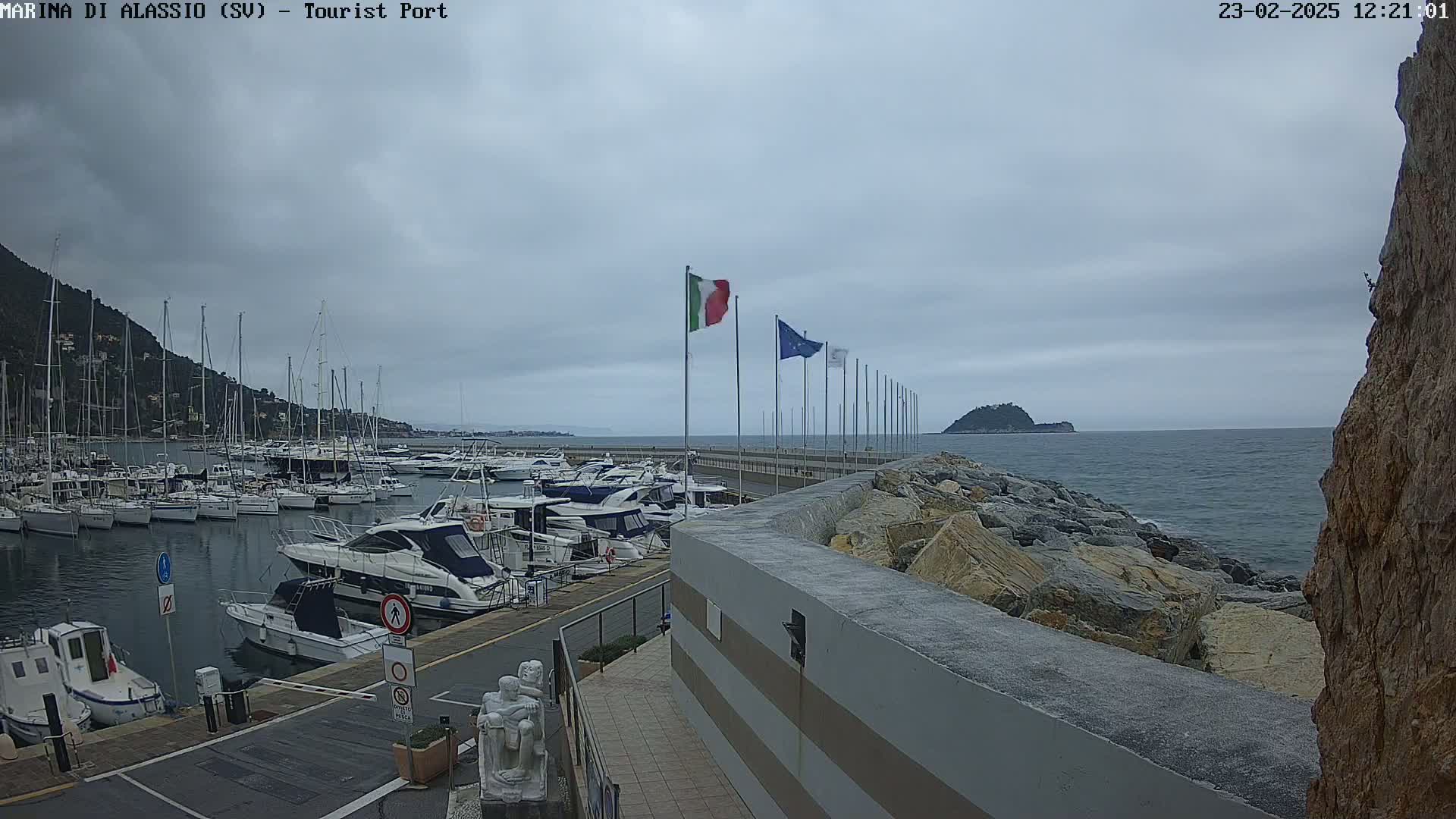 A marina filled with sailboats and motorboats is situated next to a rocky shoreline on an overcast day.
