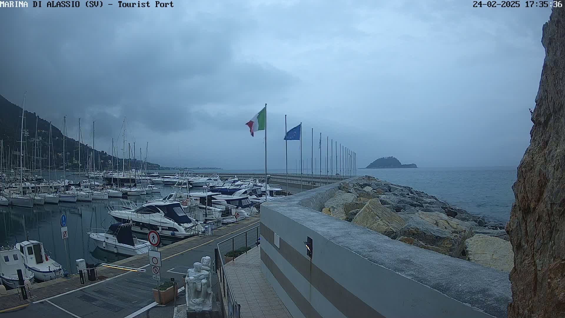 A harbor filled with numerous boats is situated under an overcast sky, with a rocky breakwater separating the marina from the sea.