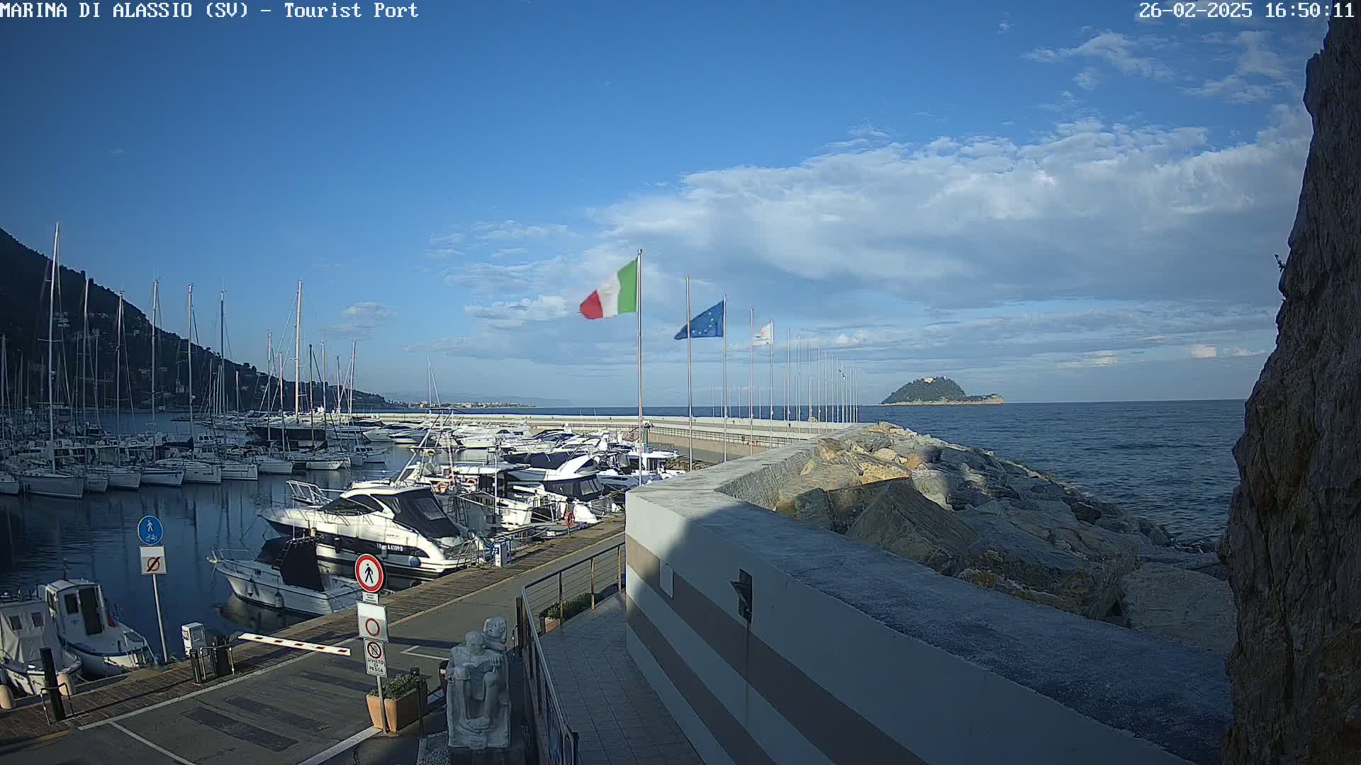 A marina filled with sailboats and motorboats is situated on a calm sea under a partly cloudy sky.