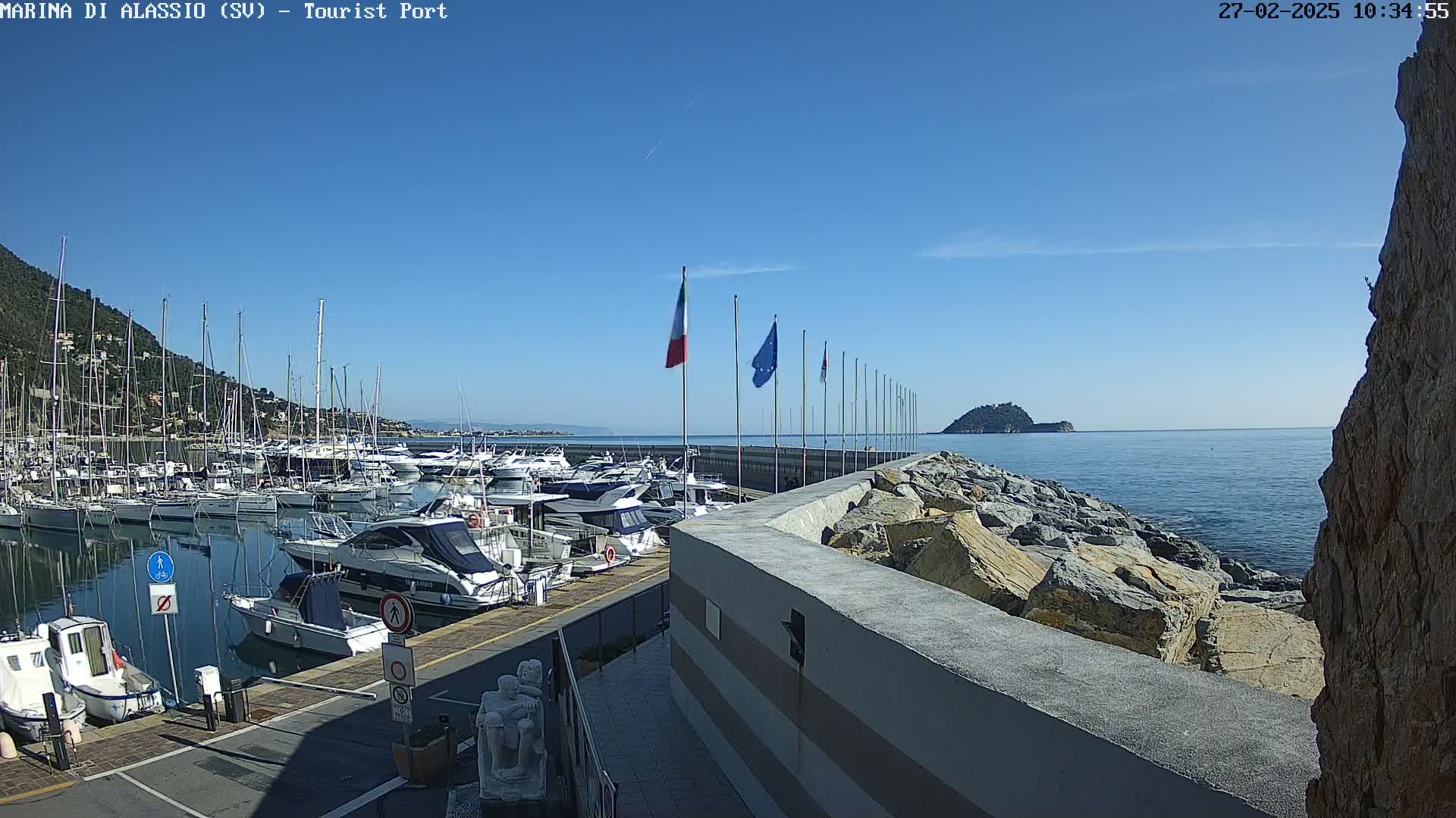 A marina filled with boats sits beside a rocky seawall on a sunny day with clear blue skies.