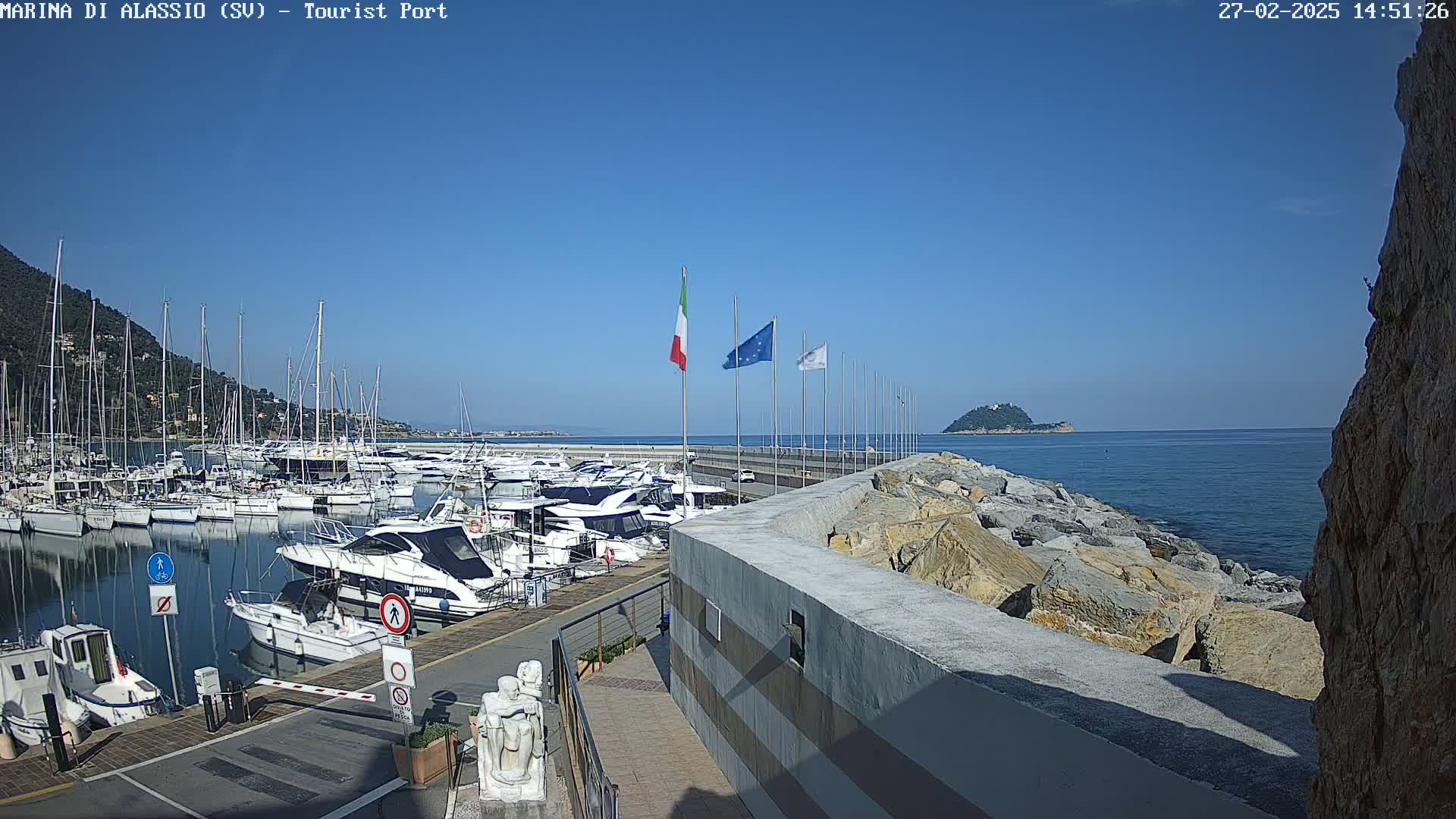 A marina filled with boats is situated on a clear, sunny day next to a rocky coastline and a small island in the distance.