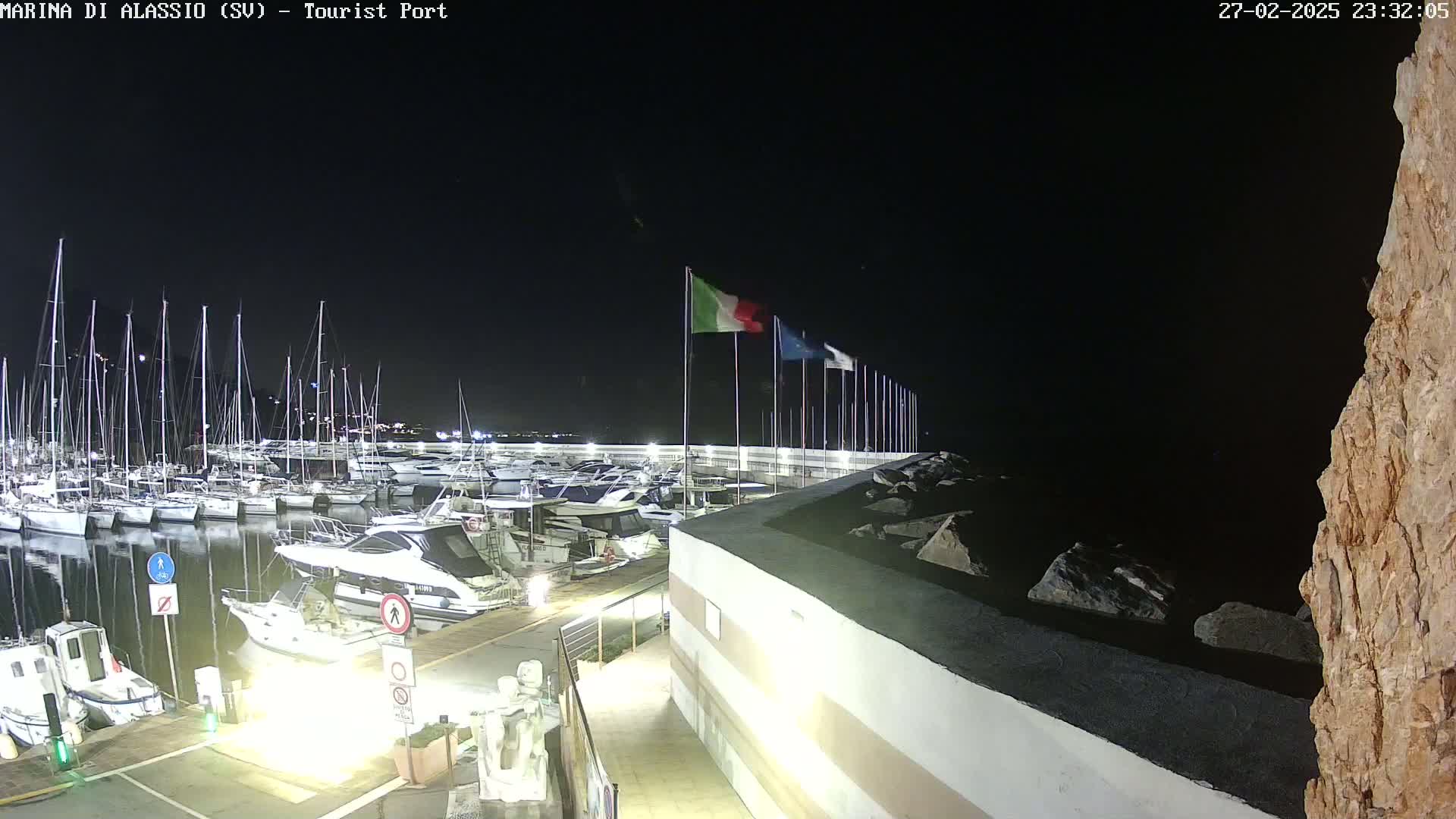 A nighttime view of a marina filled with boats, under a clear, dark sky, with Italian flags waving on a nearby wall.