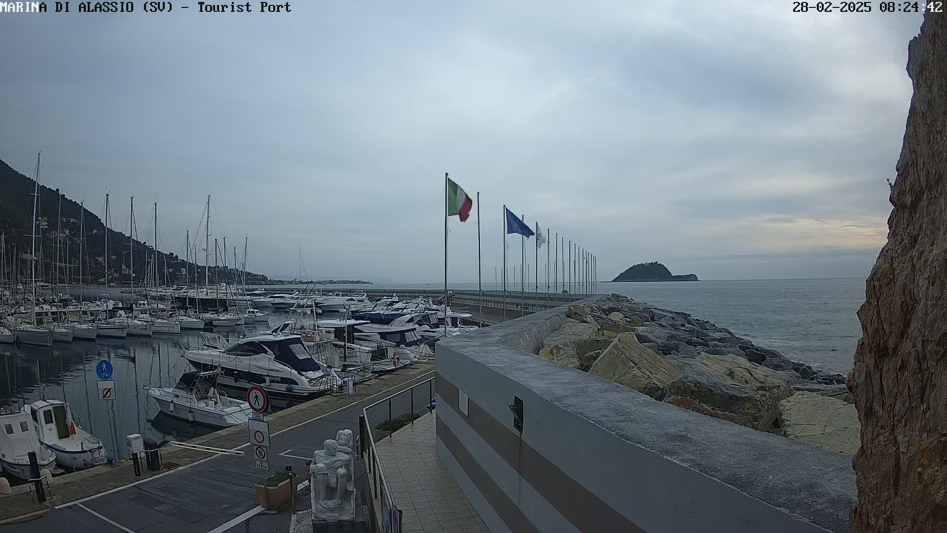 A marina filled with boats is situated next to a rocky shoreline under an overcast sky.