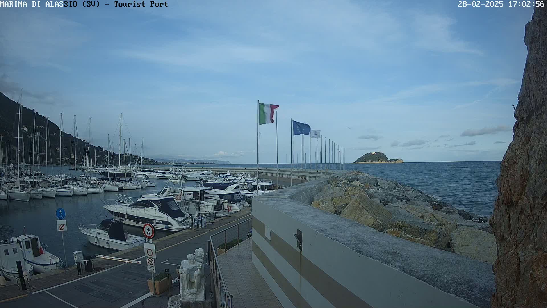 A marina filled with sailboats and motorboats is situated next to a rocky shoreline, under a mostly clear sky, with a small island visible in the distance.