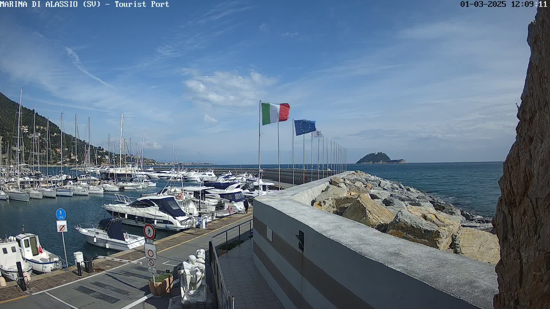 A marina filled with boats is situated next to a rocky shoreline under a mostly sunny sky.