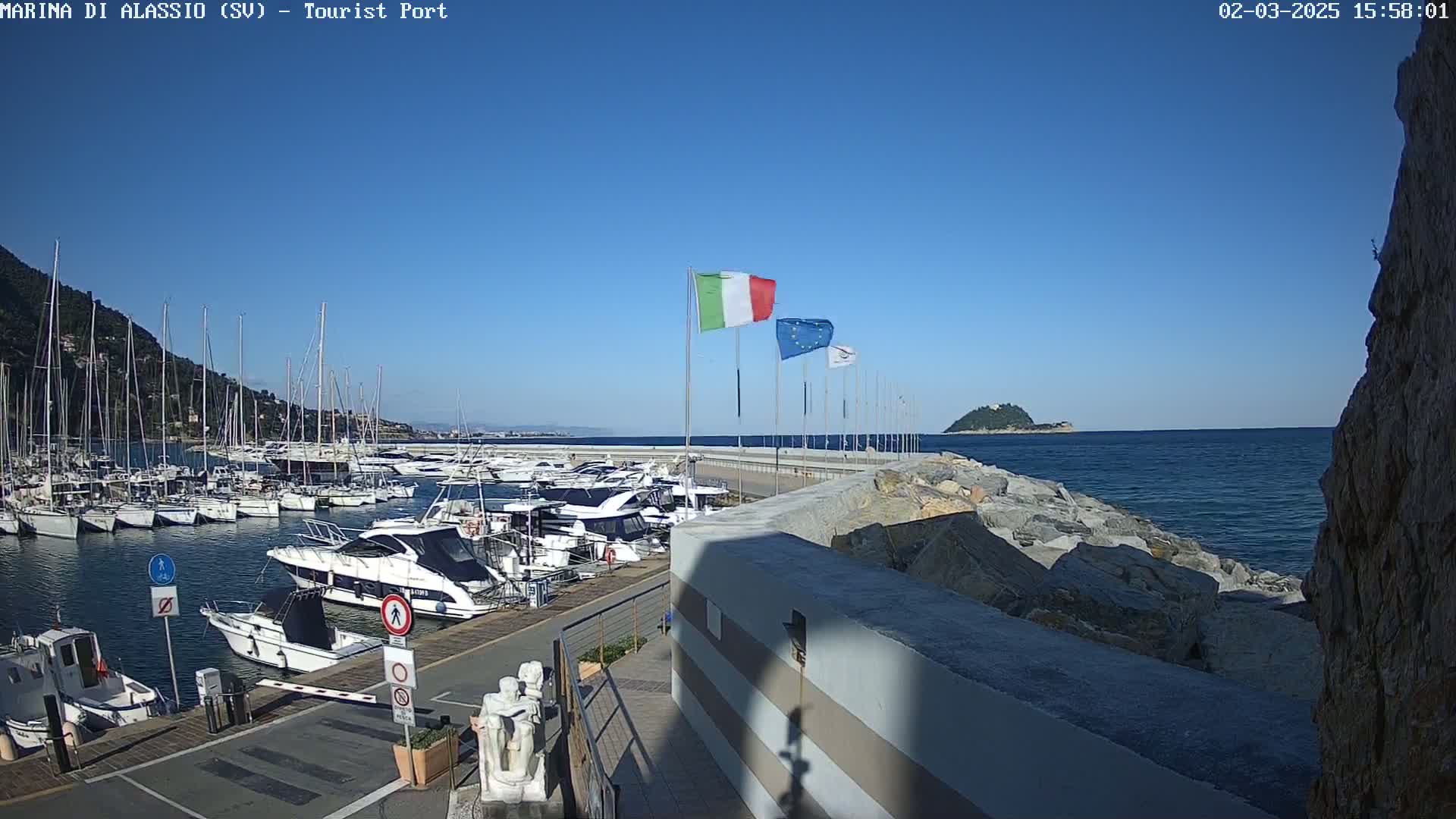 A marina filled with boats is situated next to a rocky shore on a clear, sunny day.