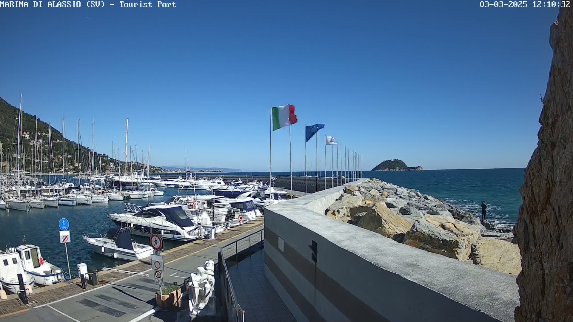 A marina filled with yachts and boats is situated on a clear, sunny day next to a rocky shoreline where a person is fishing.