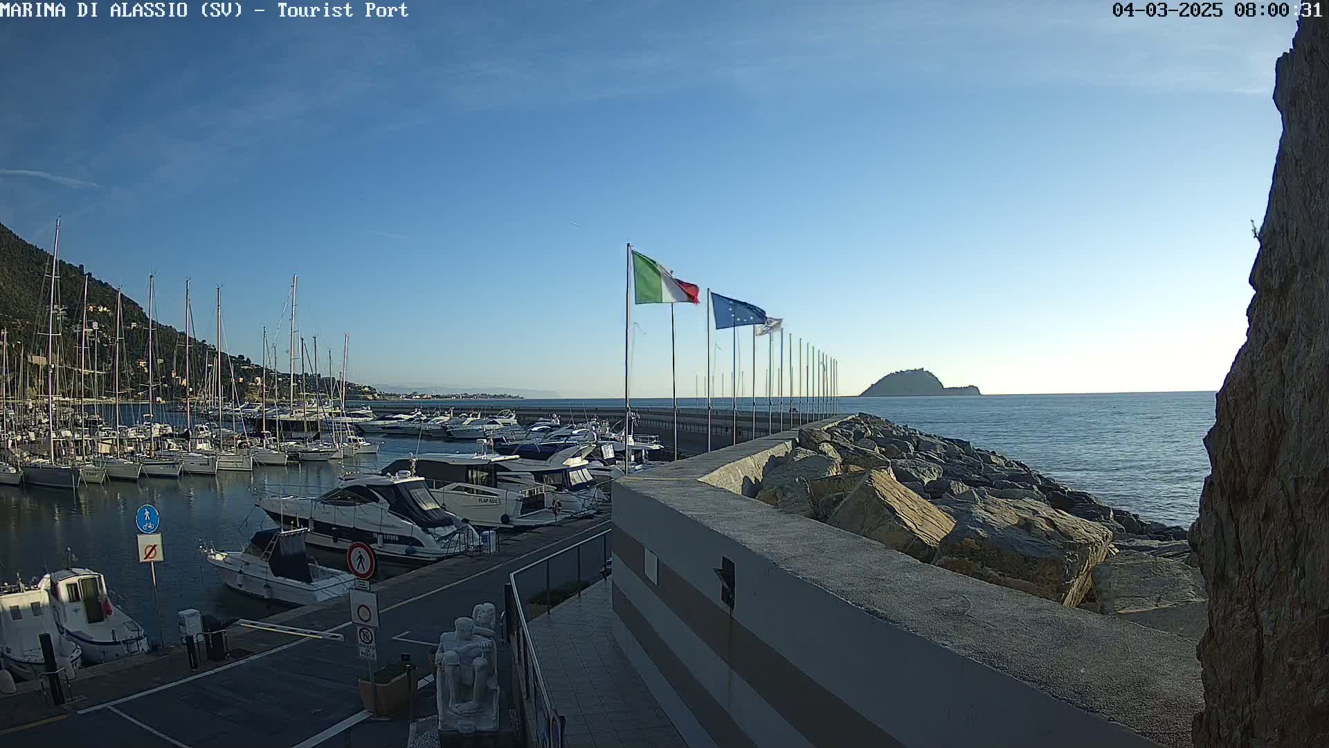 A marina filled with sailboats and motorboats is situated next to a rocky breakwater on a calm, clear day, with a small island visible in the distance.