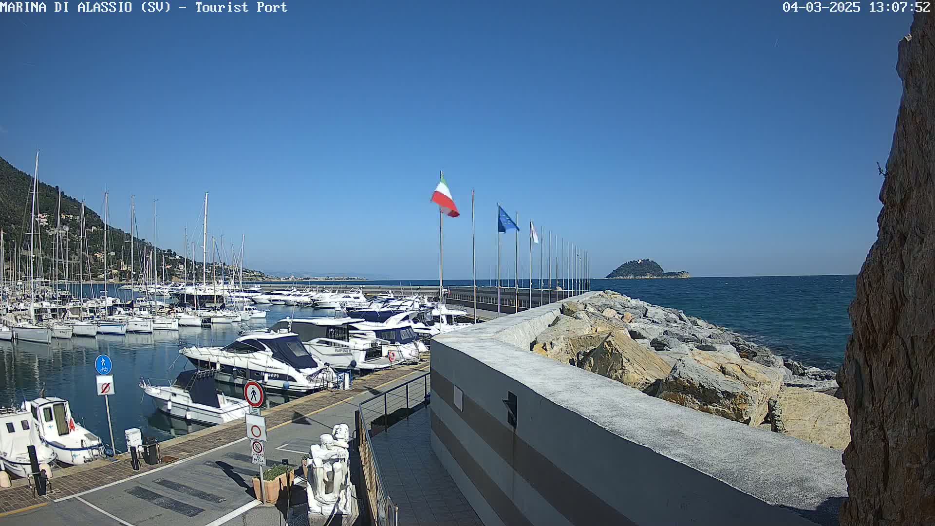 A marina filled with yachts and sailboats is situated on a calm, sunny day next to a rocky breakwater, with a small island visible in the distance.