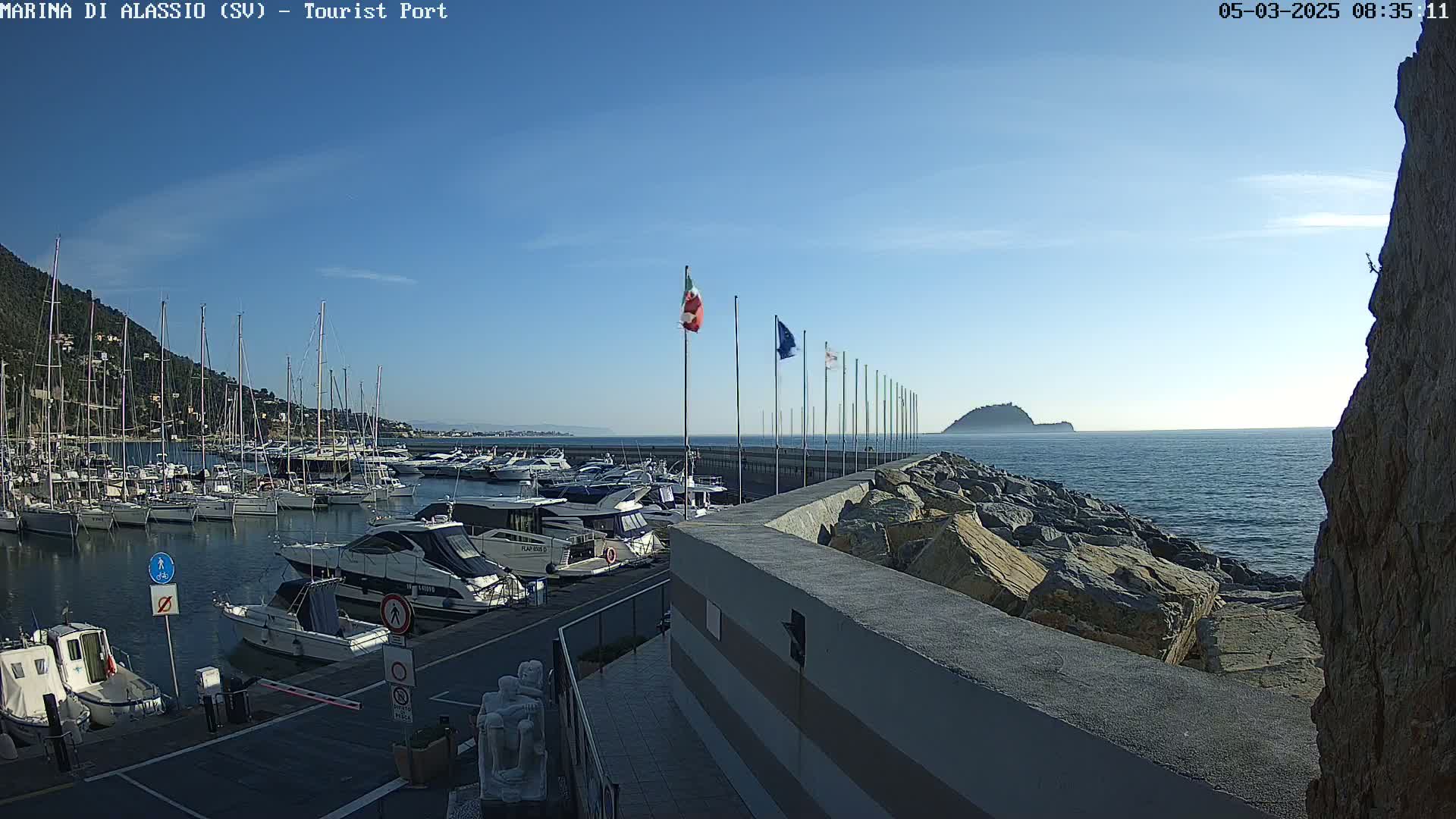 A marina filled with boats is situated next to a rocky wall, overlooking a calm sea under a clear blue sky.