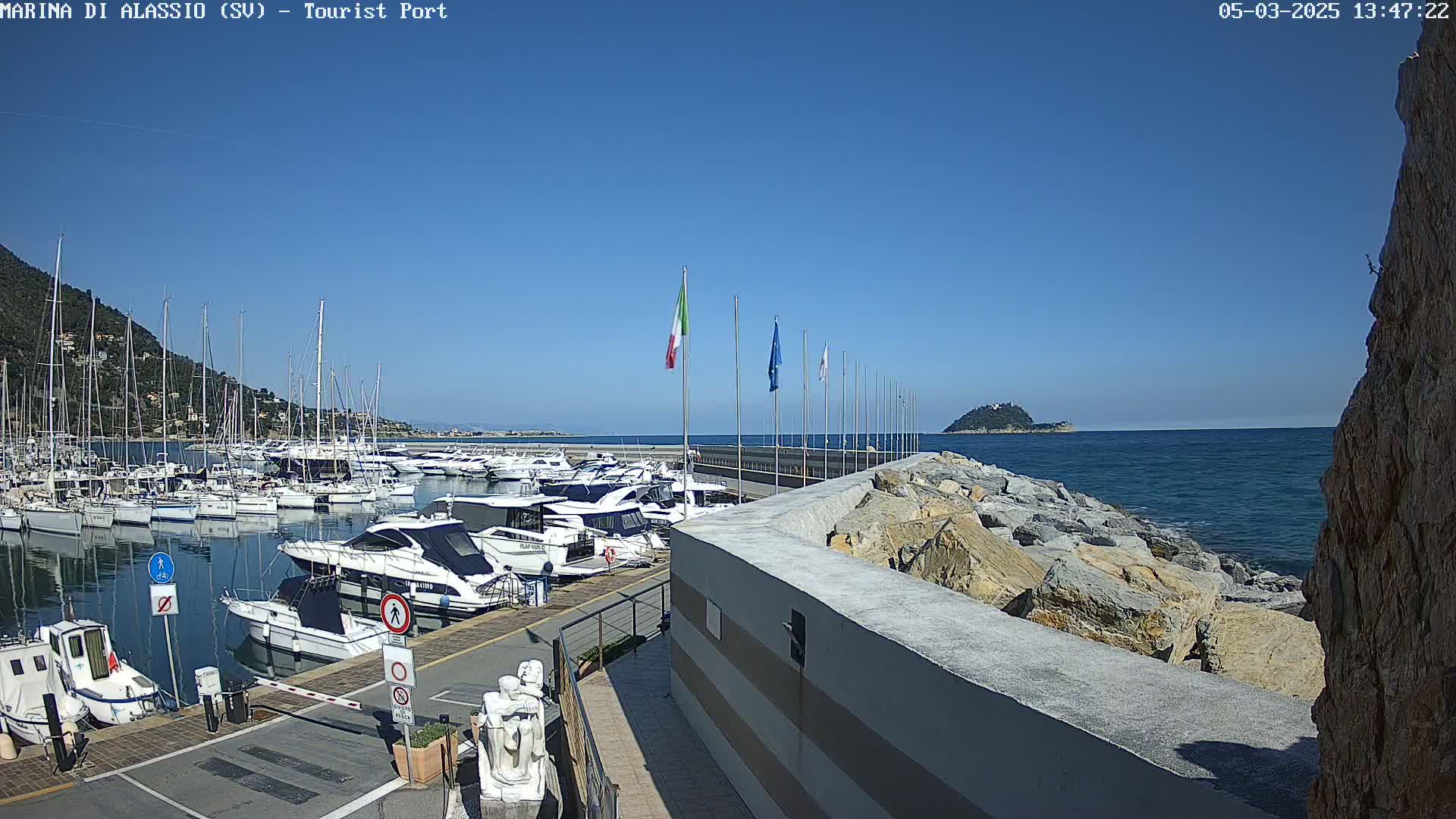 A marina filled with numerous yachts and sailboats is situated along a rocky coast under a clear blue sky.