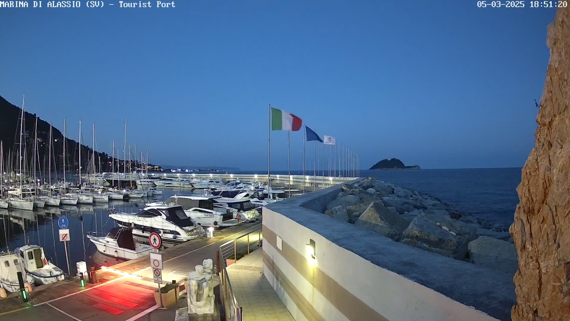 A marina filled with boats at dusk under a clear blue sky, bordered by a rocky shore.