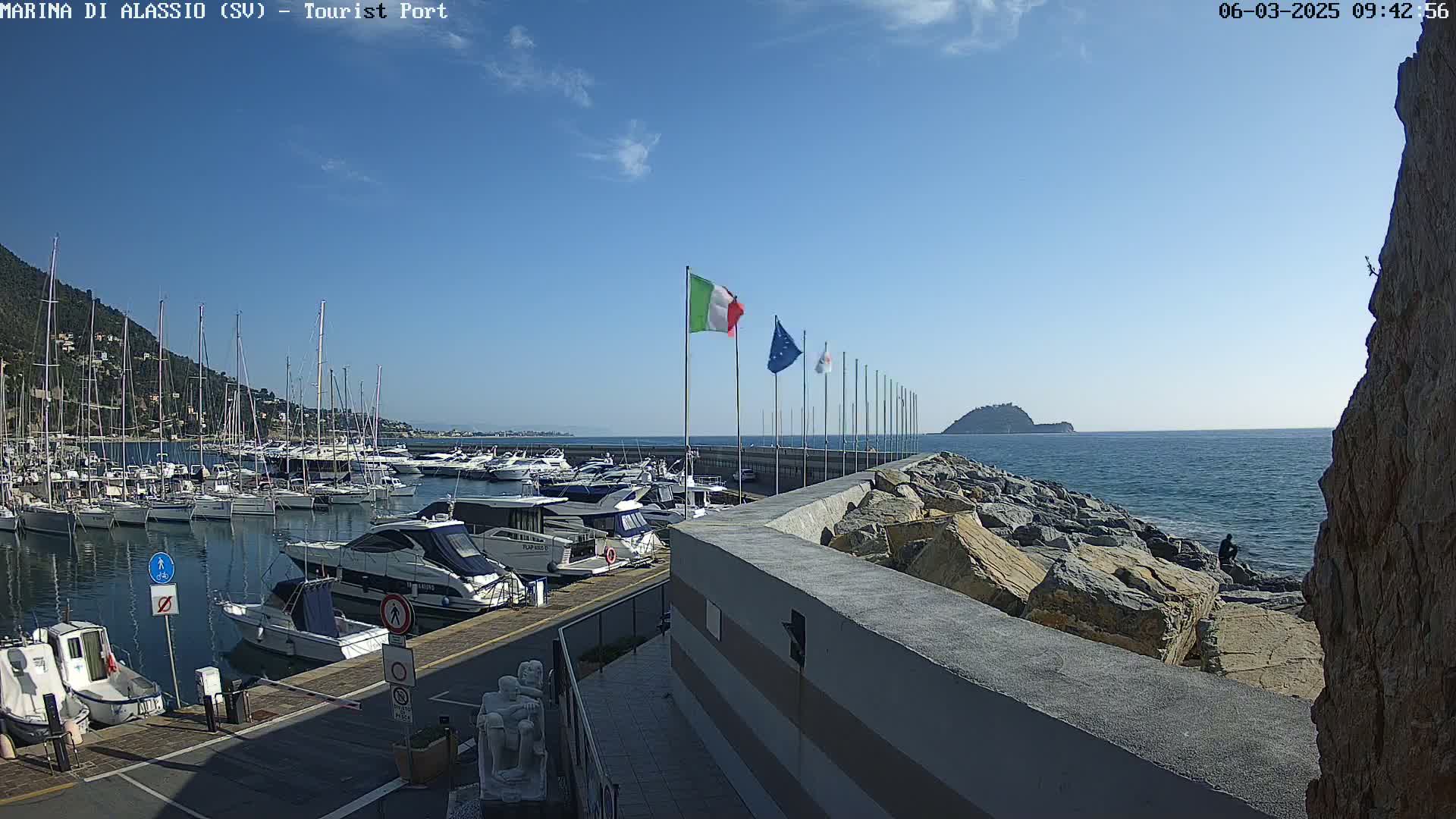 A harbor filled with numerous boats is situated next to a rocky shoreline under a clear blue sky.