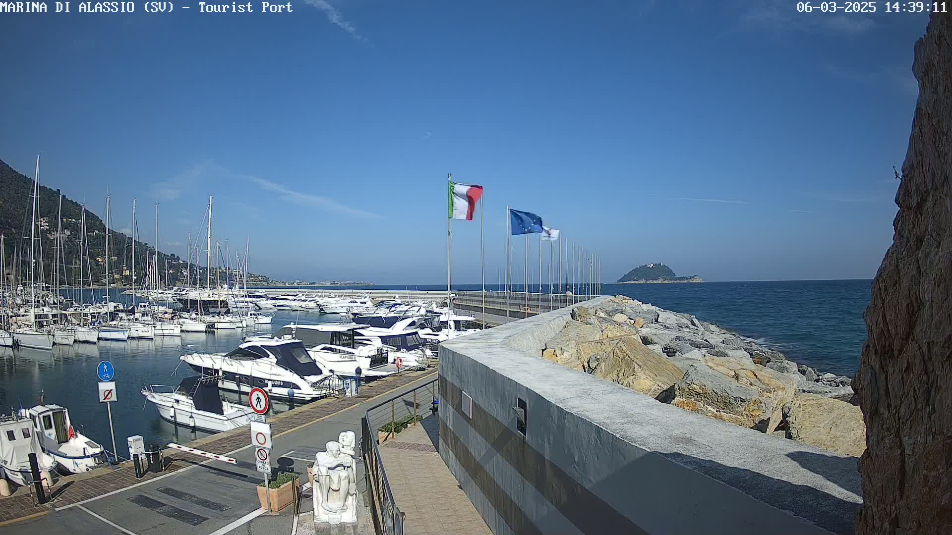 A marina filled with boats is situated beside a rocky shoreline on a clear, sunny day.