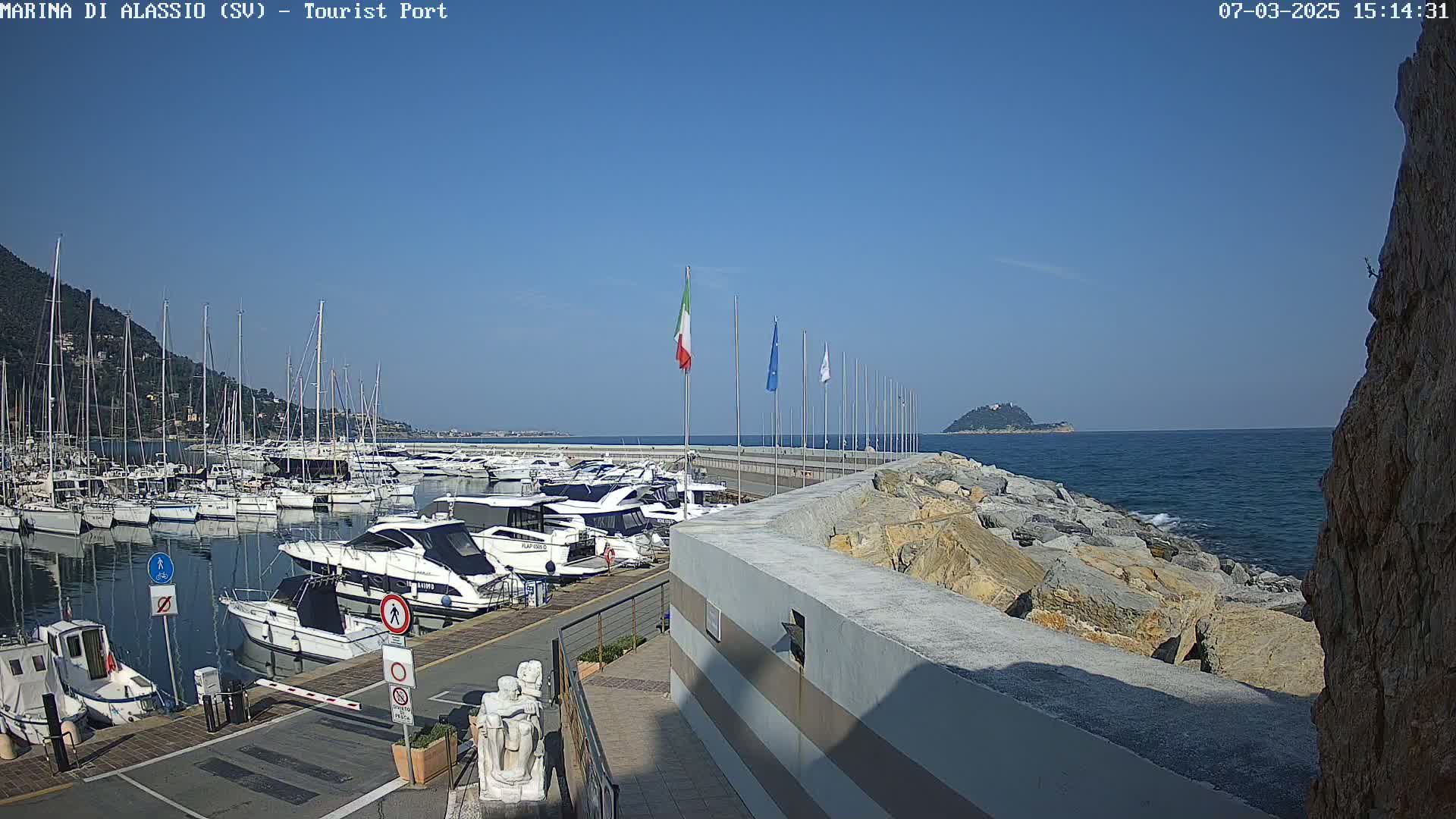 A marina filled with yachts and sailboats is situated on a calm, sunny day next to a rocky shoreline and a small island in the distance.