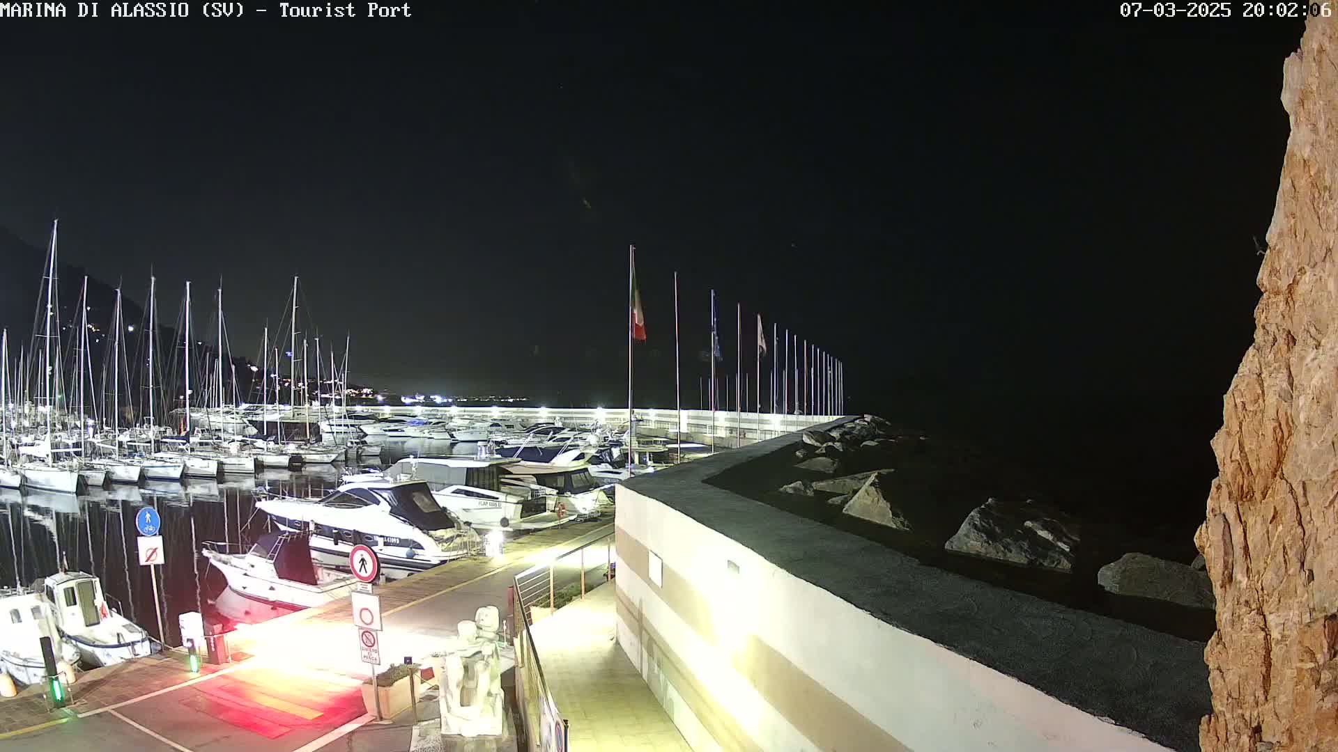 A nighttime view of a marina filled with sailboats and motorboats, docked along a wall under a clear, dark sky.