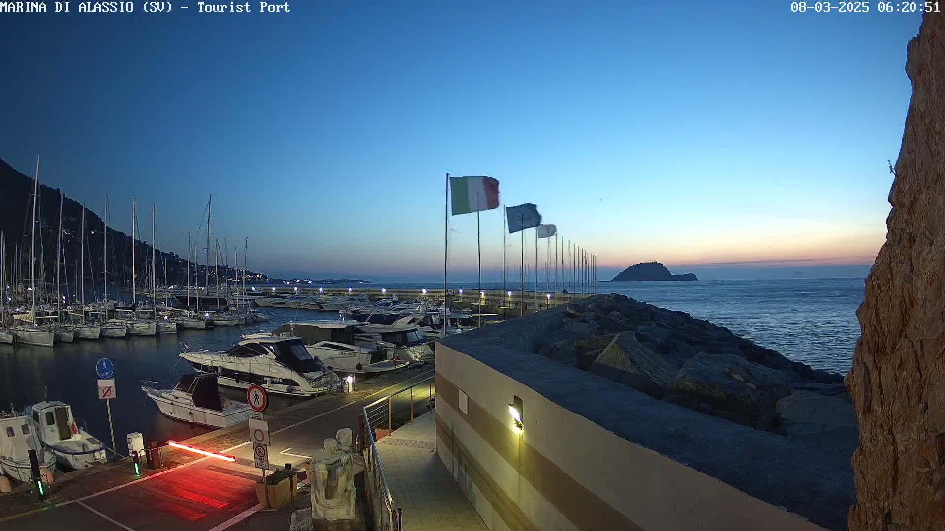 A marina filled with sailboats and motorboats at dusk under a clear, twilight sky, with a rocky breakwater and a small island visible in the distance.