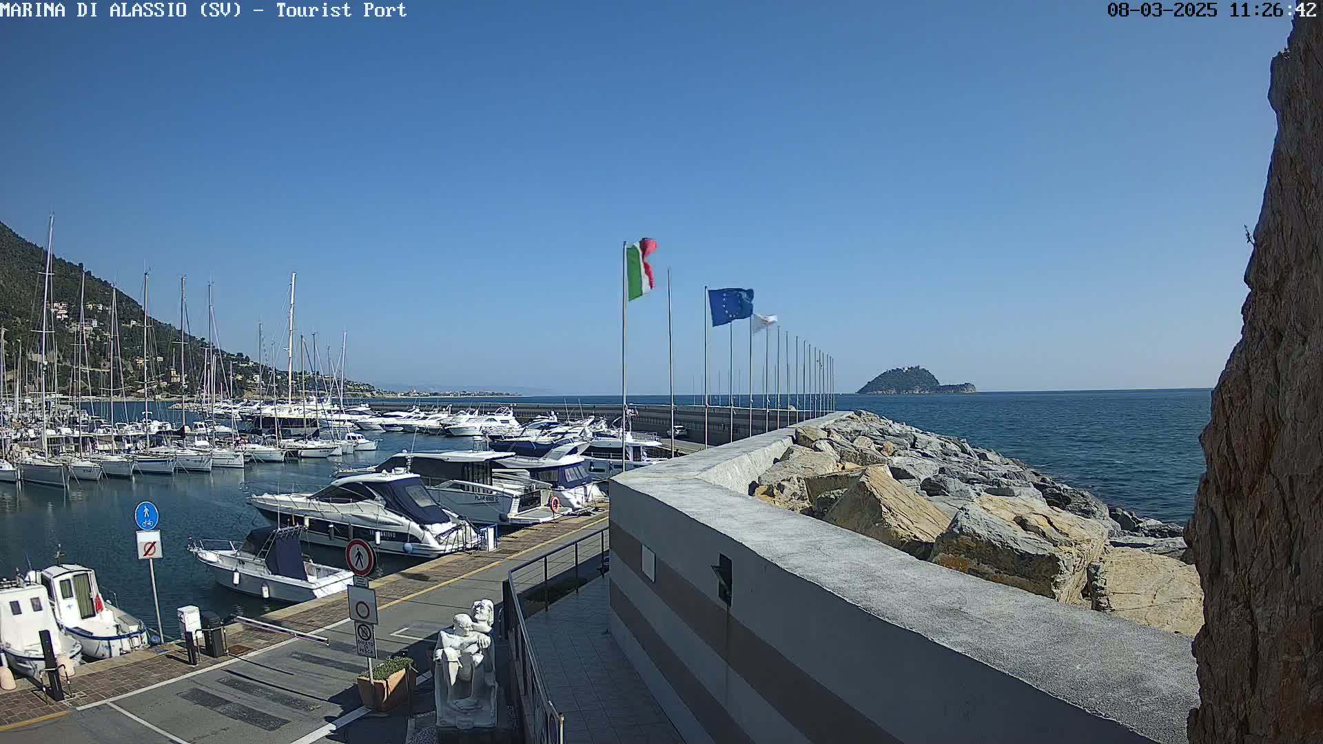 A marina filled with boats sits under a clear blue sky next to a rocky shoreline and a small island in the distance.