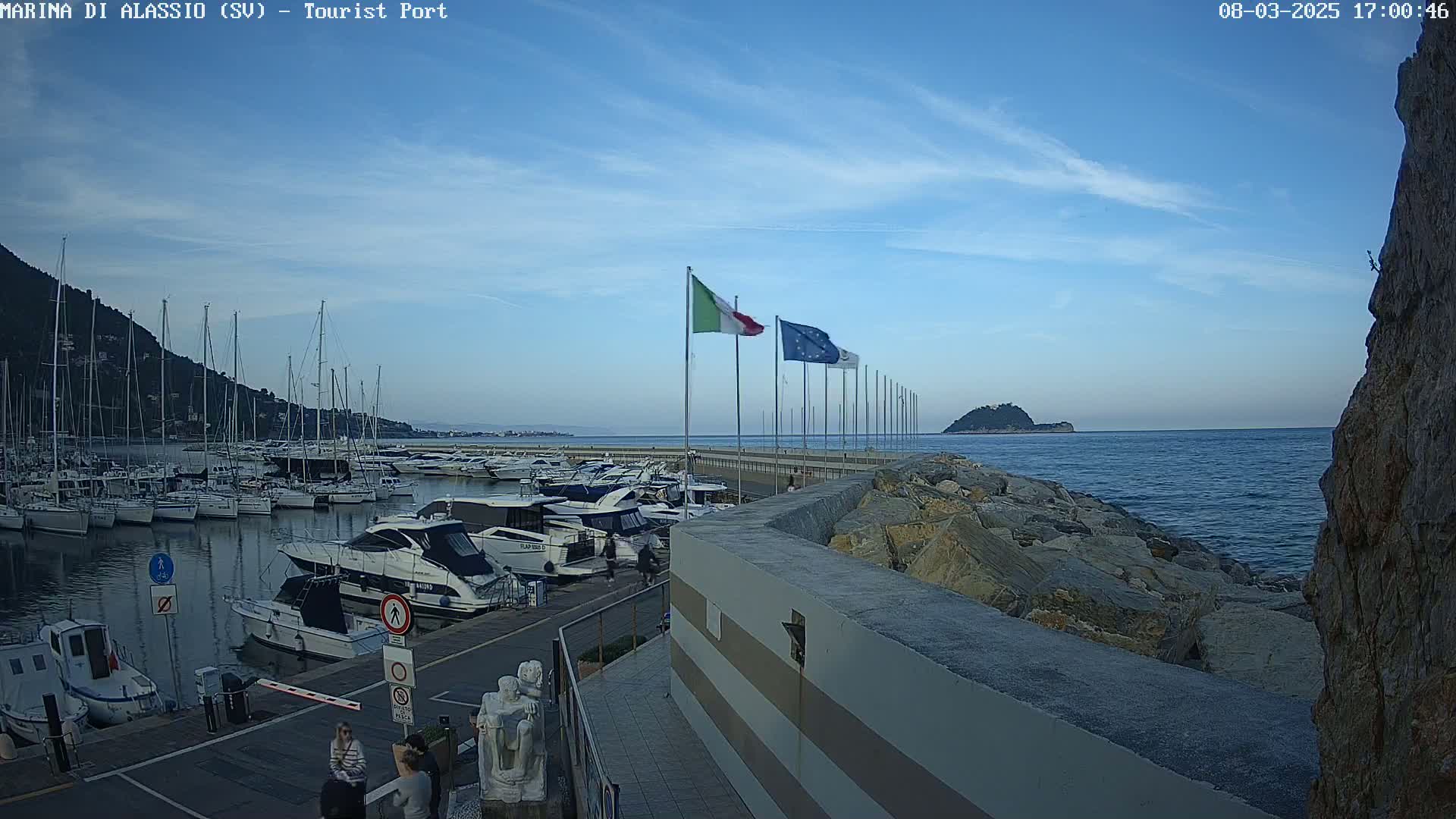A marina filled with sailboats and motorboats is situated along a rocky coastline under a clear blue sky.
