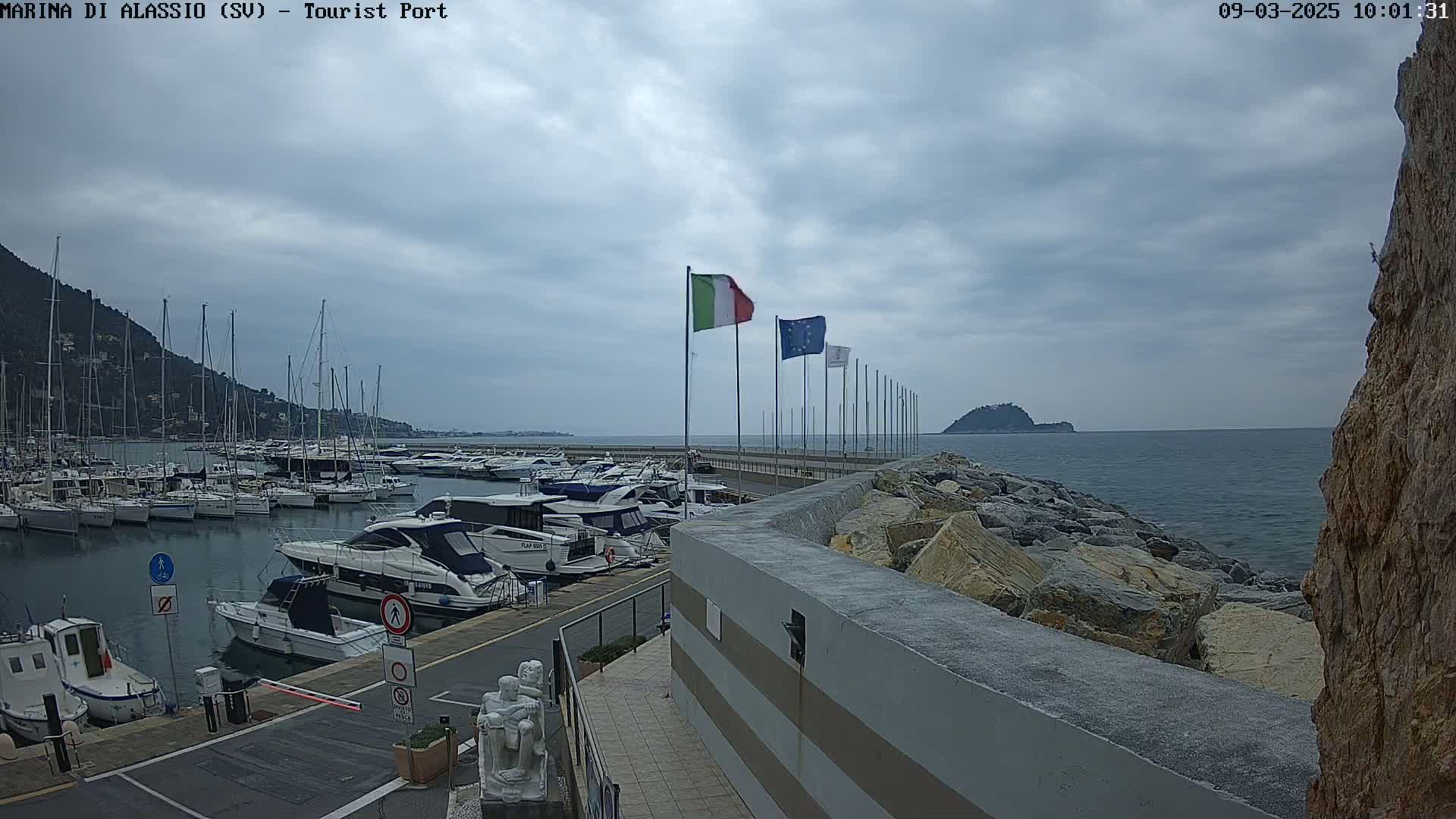 A marina filled with yachts and sailboats is situated on a calm, overcast day next to a rocky breakwater that extends into a body of water toward a small island.