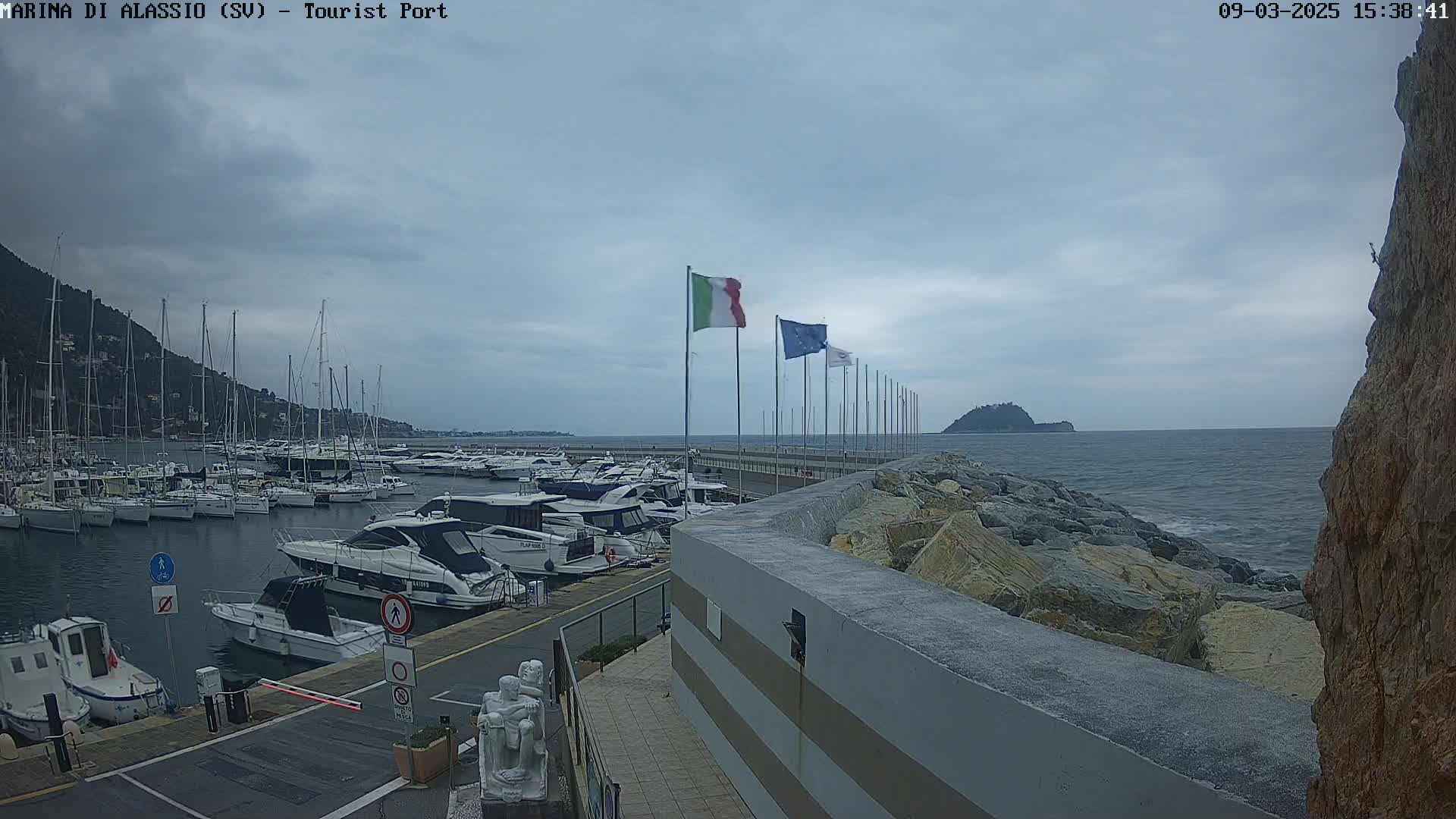 A harbor filled with sailboats and motorboats is seen under an overcast sky, with a rocky shoreline and calm water in the background.