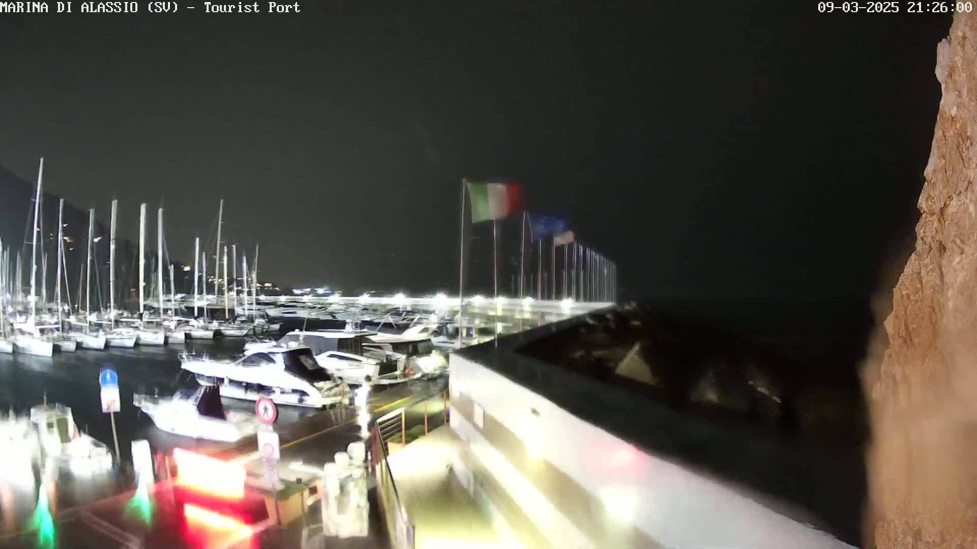 A nighttime view of a marina filled with boats, under a dark sky, with Italian flags waving in the wind.