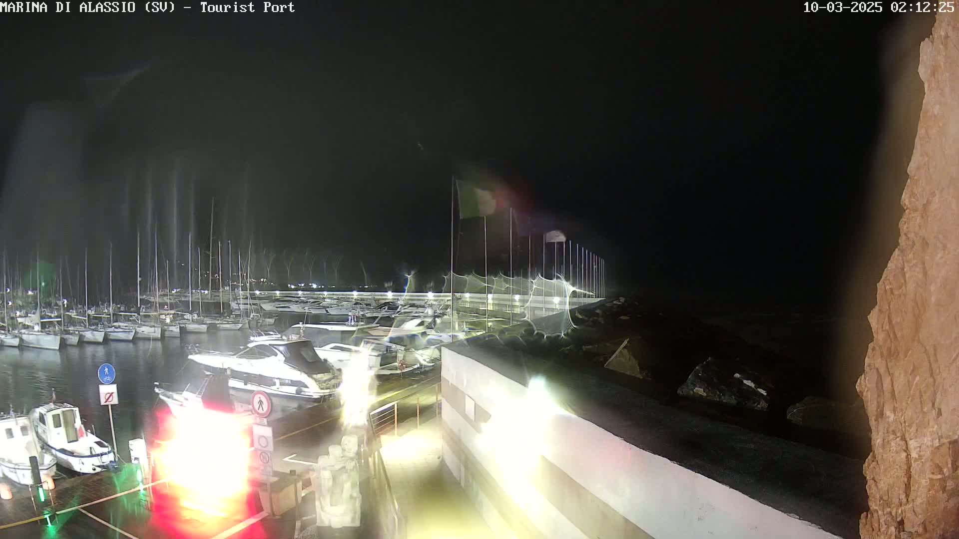 A nighttime view of a marina with numerous boats docked, under a dark sky with rain.