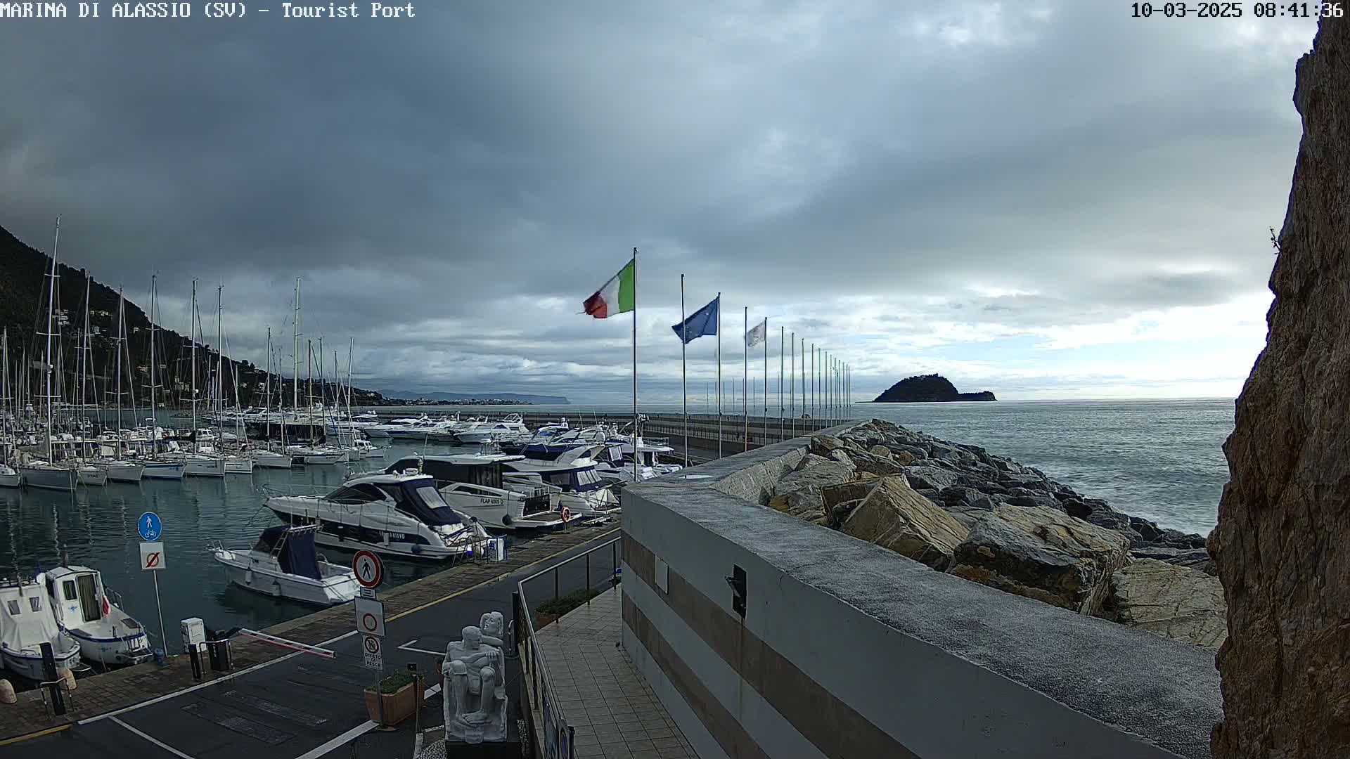 A marina filled with yachts and sailboats is situated next to a rocky breakwater under a cloudy sky, with calm to moderately choppy water visible beyond.